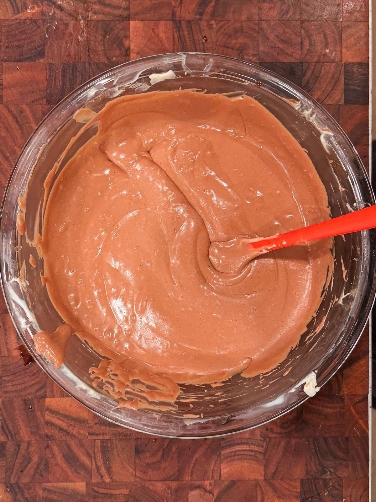 A glass bowl filled with creamy, light brown chocolate mascarpone cream is being mixed with a red spatula, resting on a wooden surface.