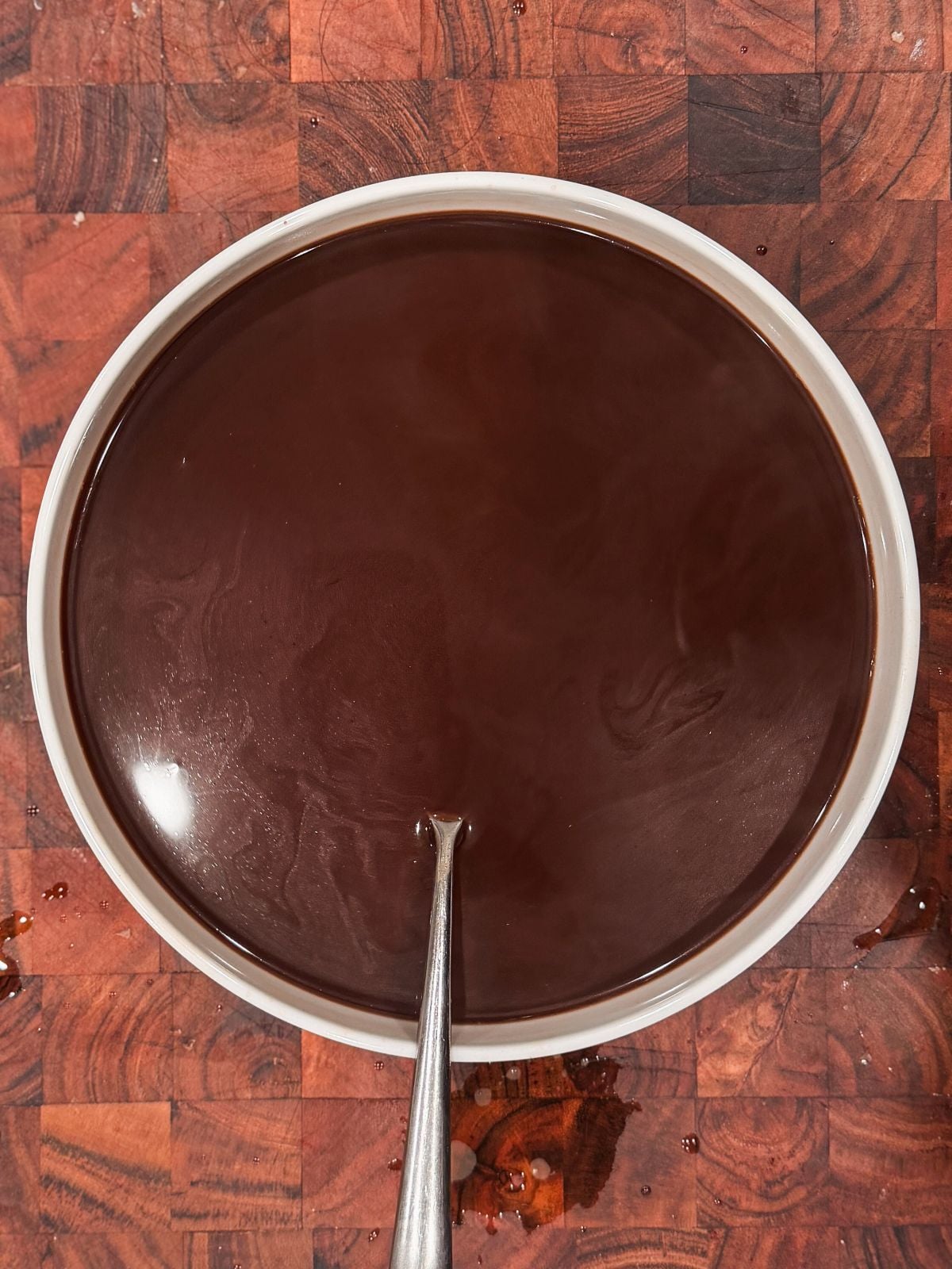 A white bowl filled with glossy, dark chocolate coffee liquid sits on a wooden surface, with a metal spoon resting inside the bowl.