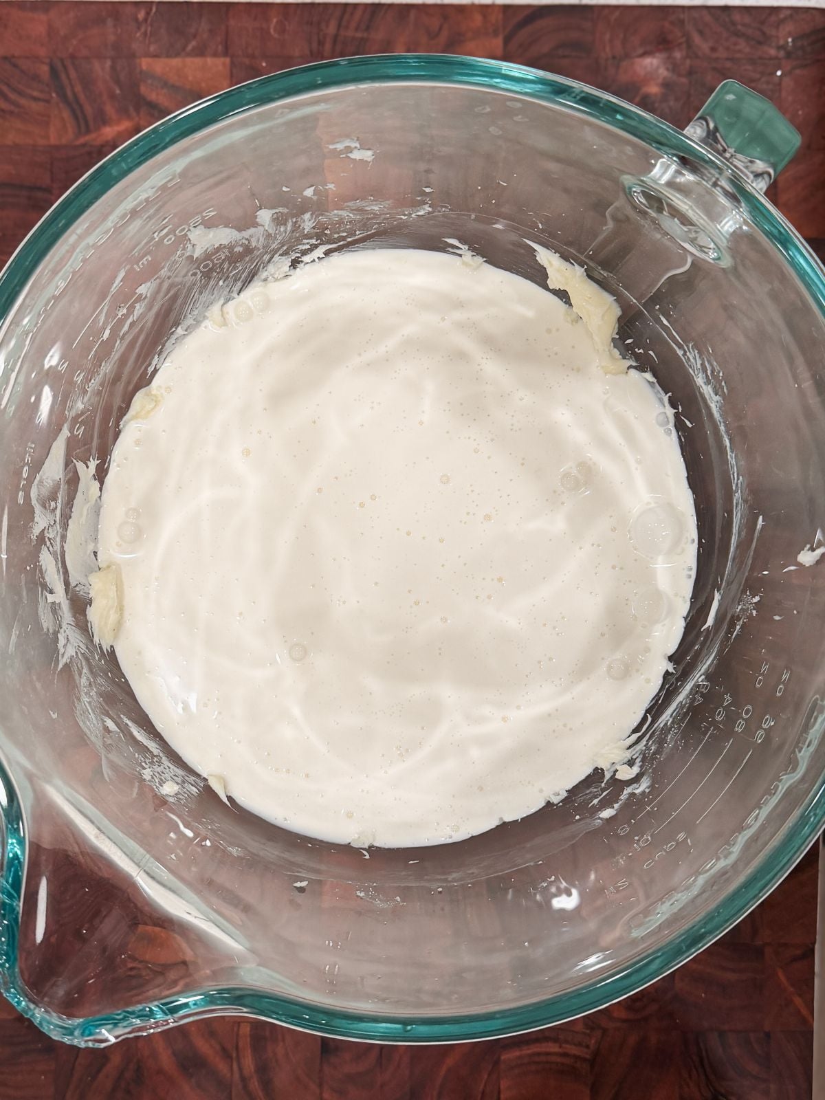 A glass mixing bowl filled with white cream sits on a wooden countertop. The sides of the bowl have some batter residue.