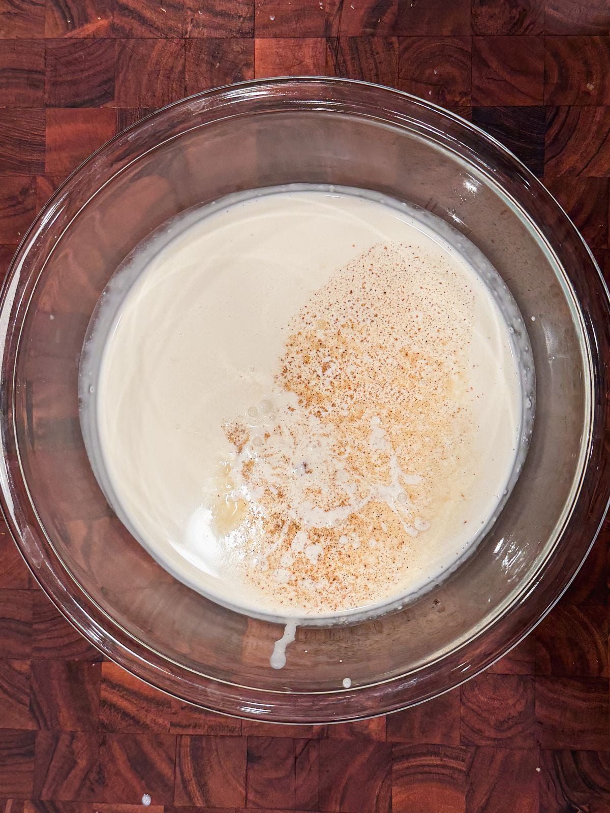 A clear glass bowl on a wooden surface contains a cream, sugar and cardamom powder on top.