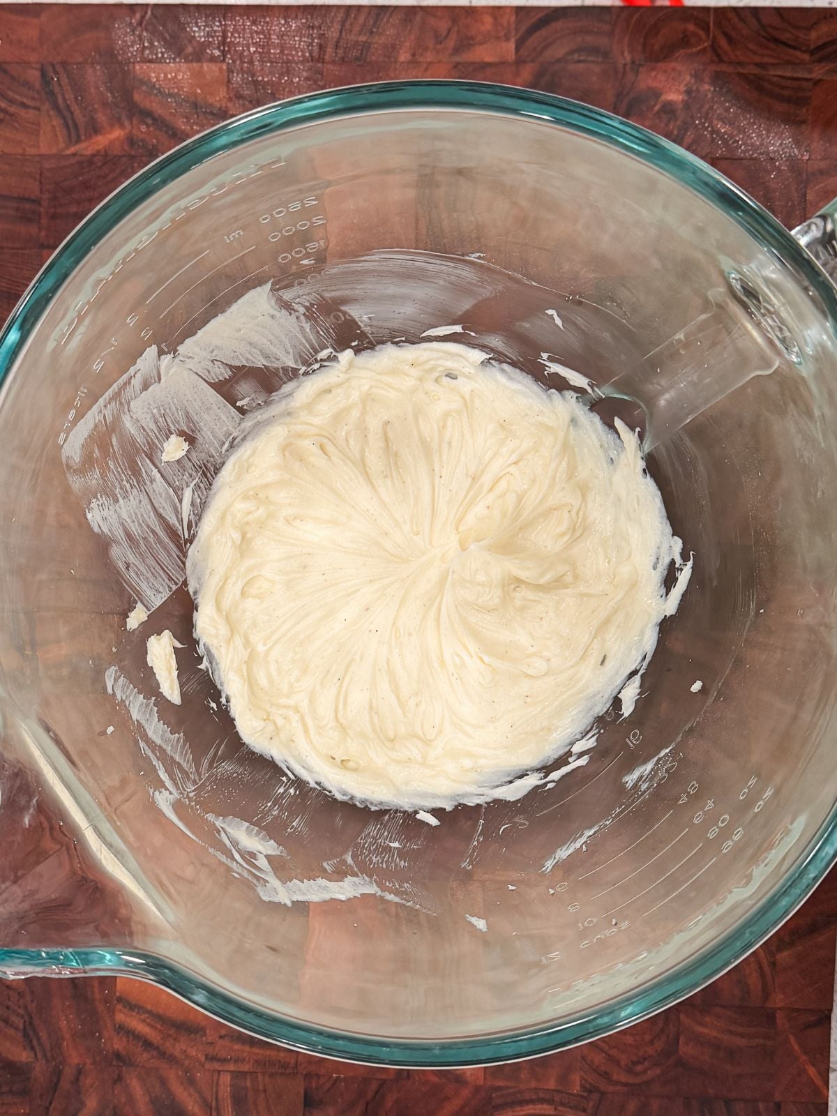 A clear glass mixing bowl containing a small amount of creamy, off-white whipped cream cheese, swirled in the center, sits on a dark wooden surface.
