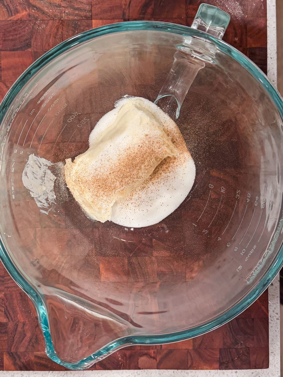 A glass mixing bowl on a wooden surface contains cream cheese, sugar, and sprinkled cardamom, ready to be mixed.