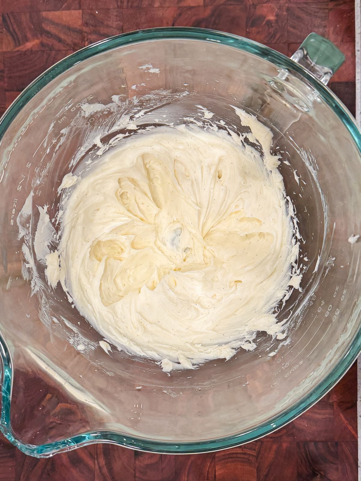 A glass mixing bowl containing smooth, creamy mixture of cream cheese and sugar sits on a dark wooden surface.