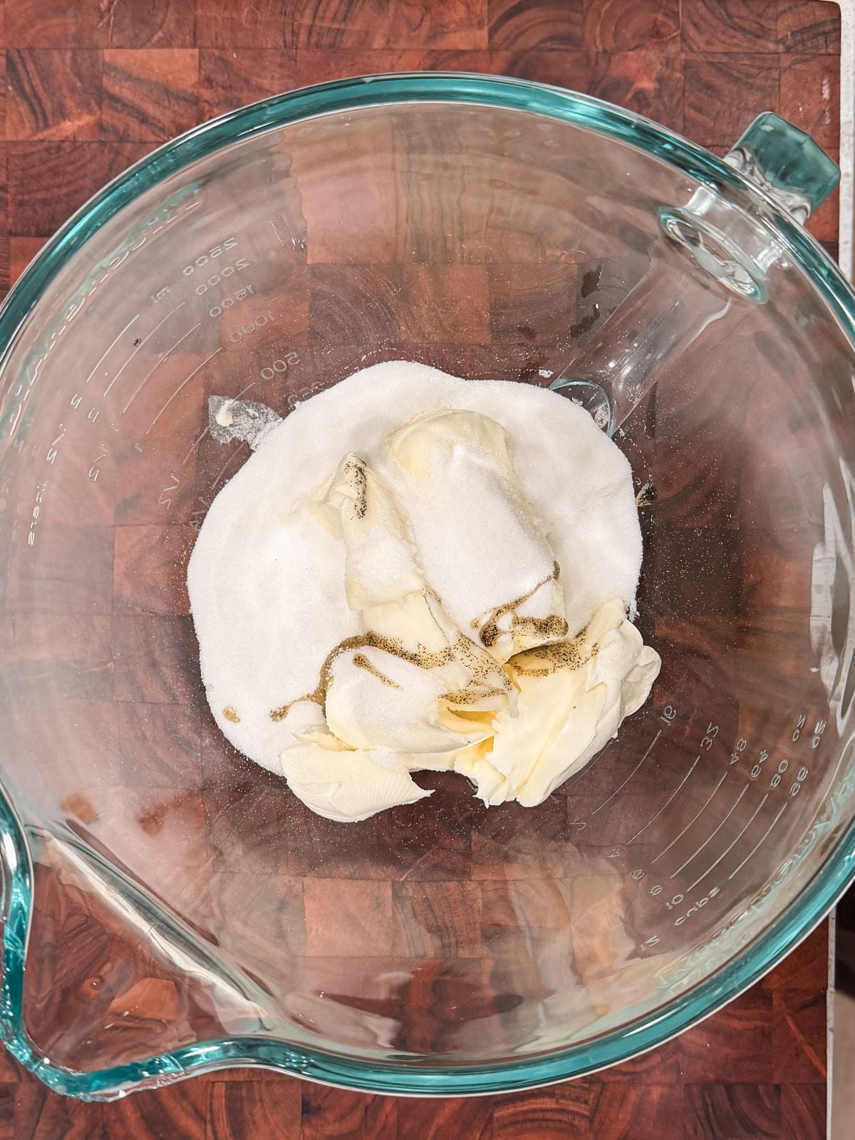 A glass mixing bowl on a wooden surface contains cream cheese, granulated sugar, and vanilla extract, ready to be mixed together.