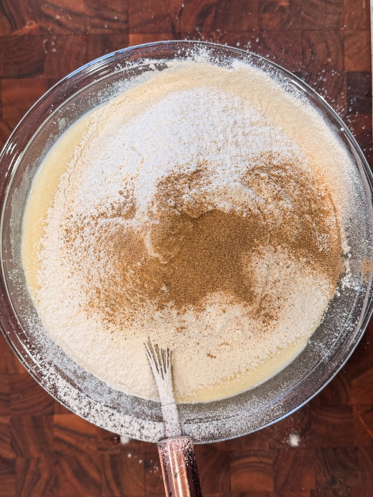A glass bowl on a wooden surface contains flour, sugar, and cardamom on top of a wet batter, with a metal whisk partially submerged in the mixture, ready for mixing.