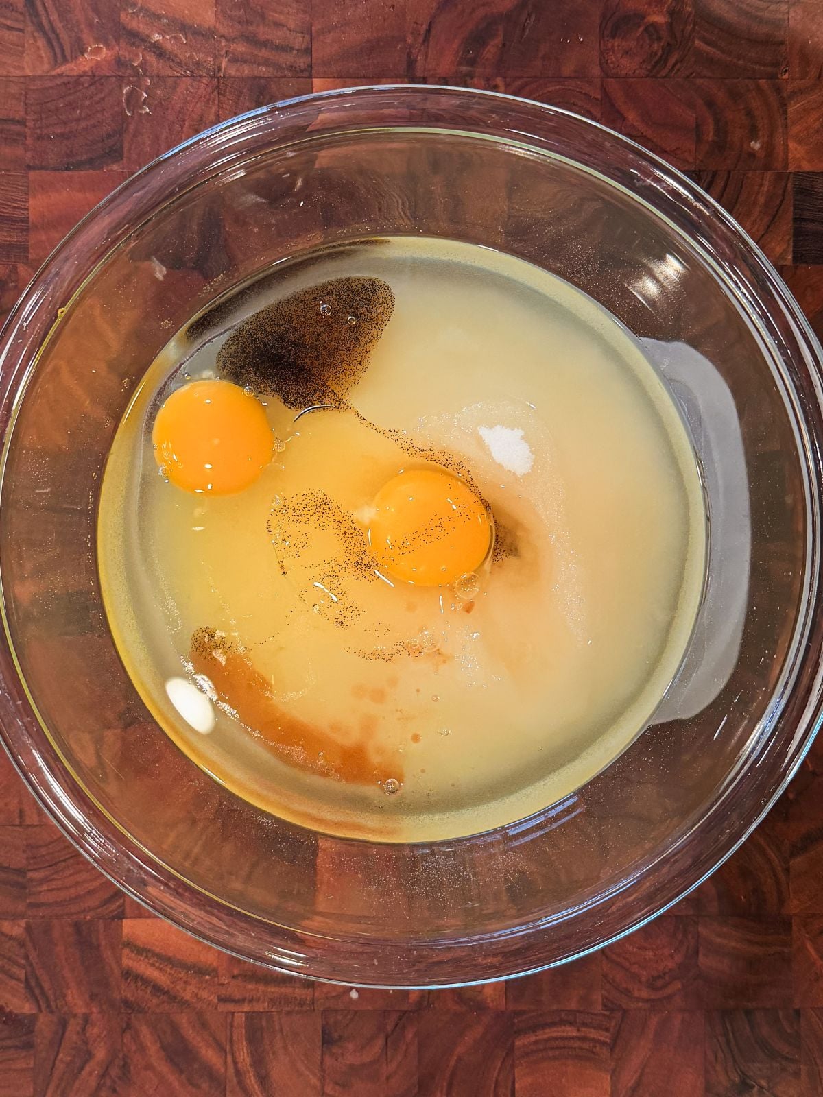 A glass bowl on a wooden surface containing two cracked eggs, sugar, vanilla extract, and other wet ingredients, ready to be mixed.