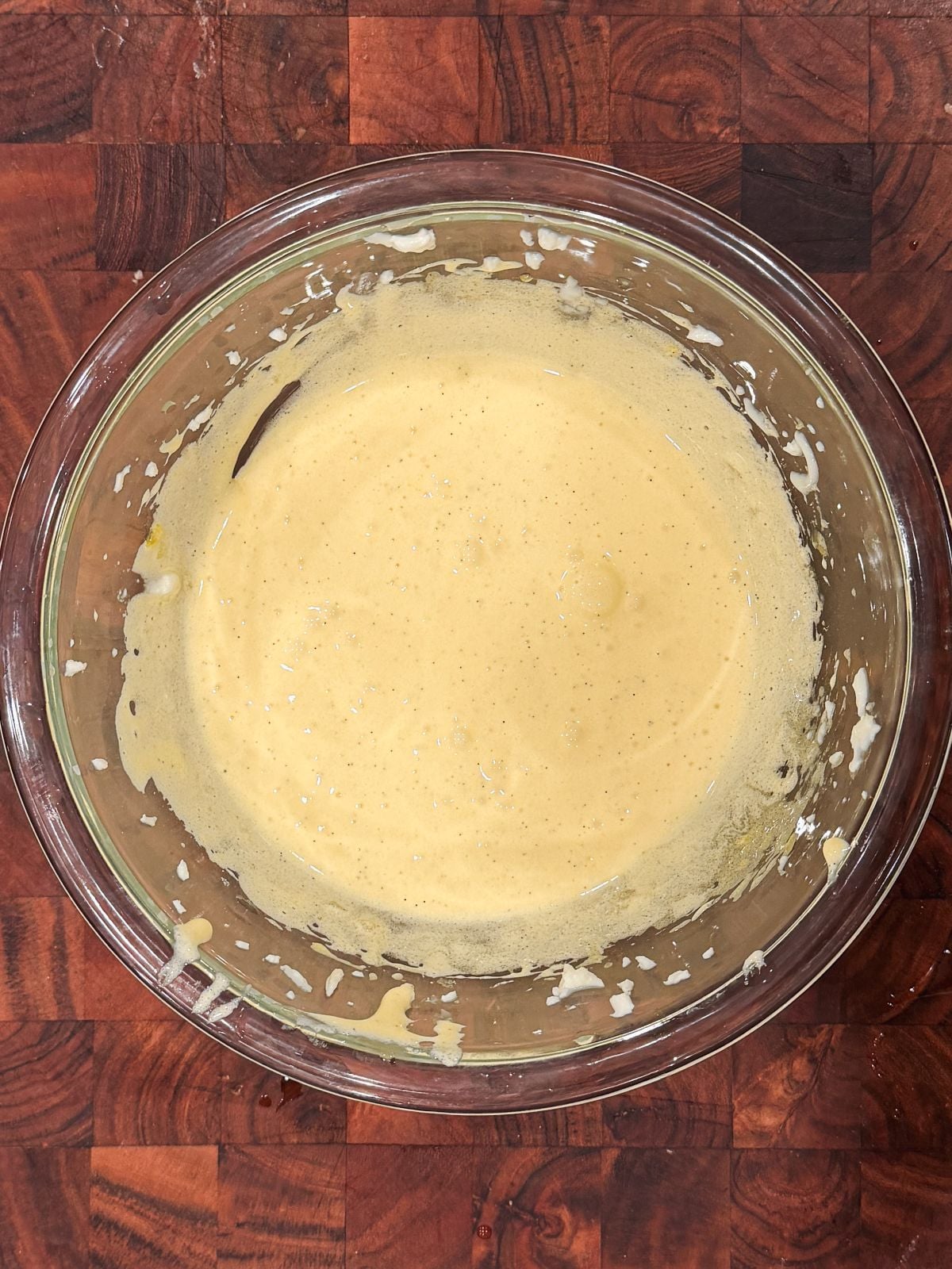 A glass mixing bowl filled with a pale yellow mascarpone mixture sits on a wooden surface. Some batter is splattered along the sides of the bowl.