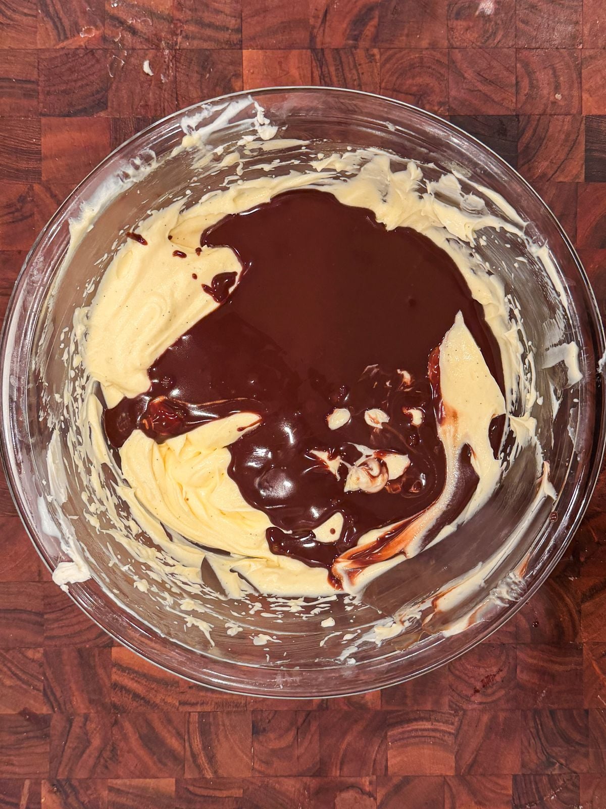 A glass bowl on a wooden surface contains yellow mascarpone cream with chocolate ganache poured on top, partially mixing but not yet fully combined.