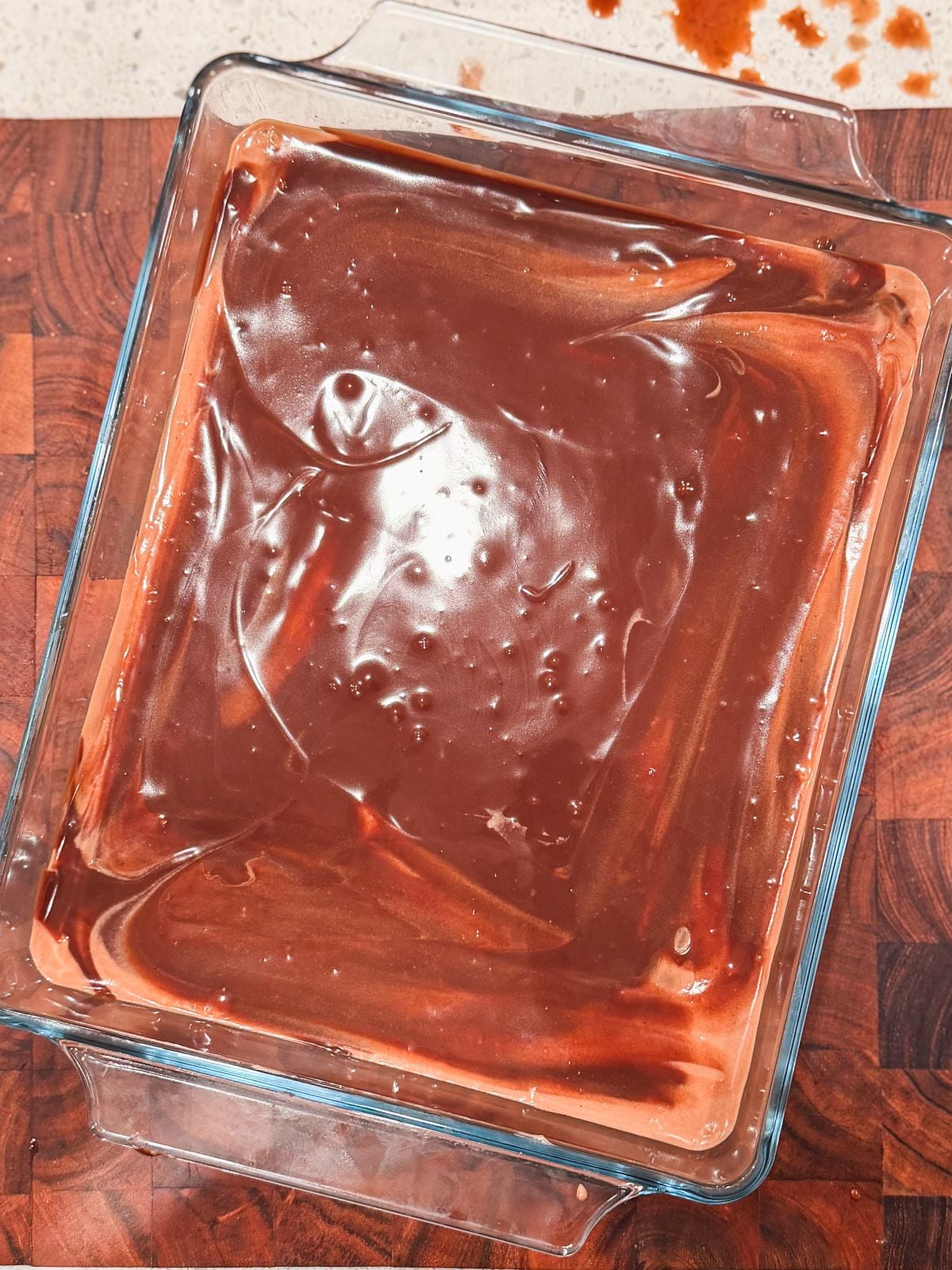 A glass baking dish filled with a smooth, glossy layer of chocolate ganache topping sits on a wooden surface, with a few chocolate smudges visible on the counter nearby.