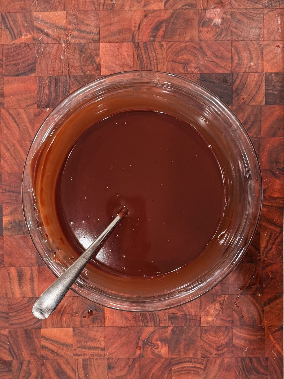 A glass bowl filled with smooth, glossy chocolate ganache, with a metal spoon resting in it, sits on a dark wooden, patterned countertop.