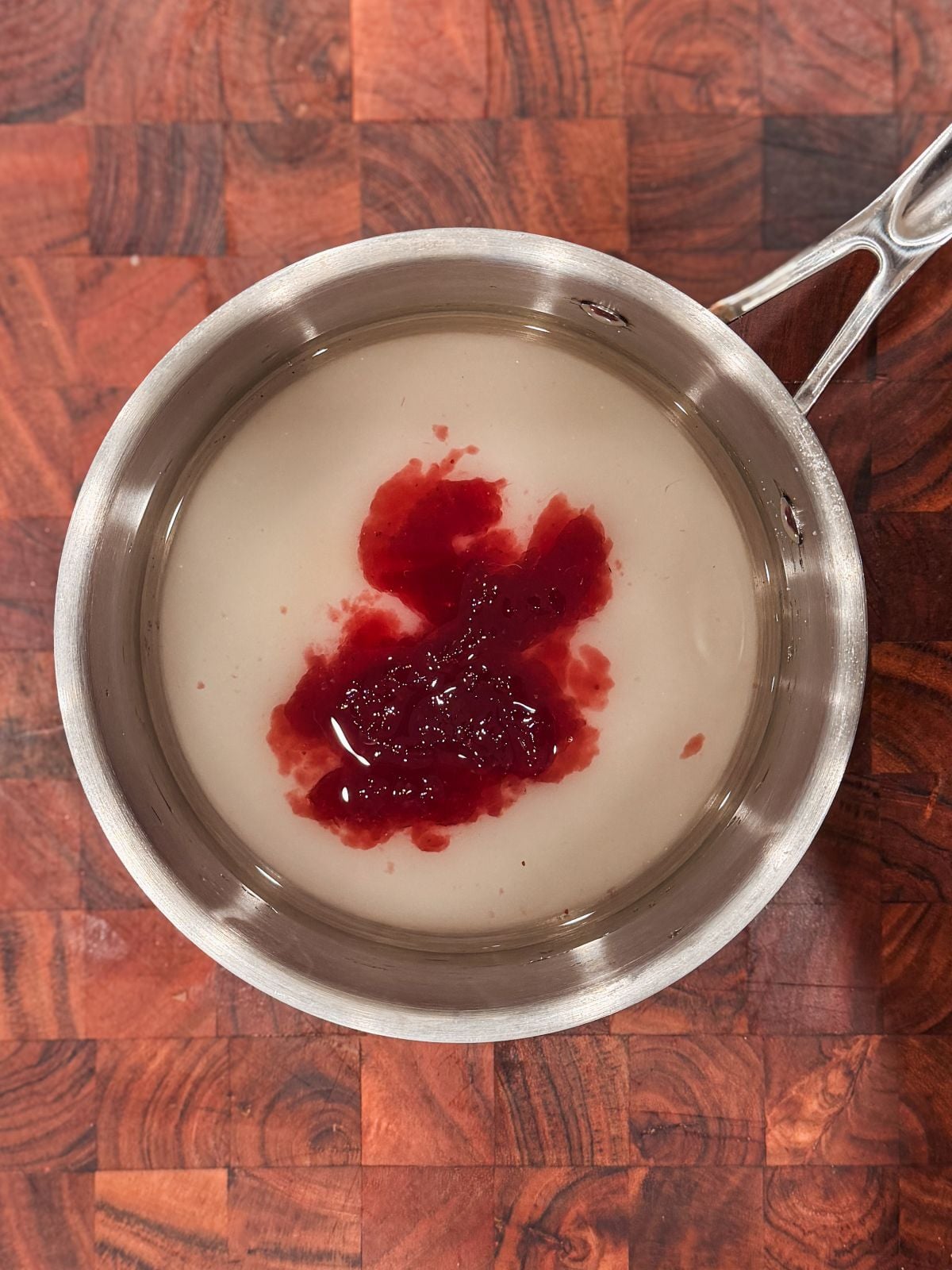 A metal saucepan containing water, sugar and jam in the center, placed on a wooden surface with a checkered pattern.
