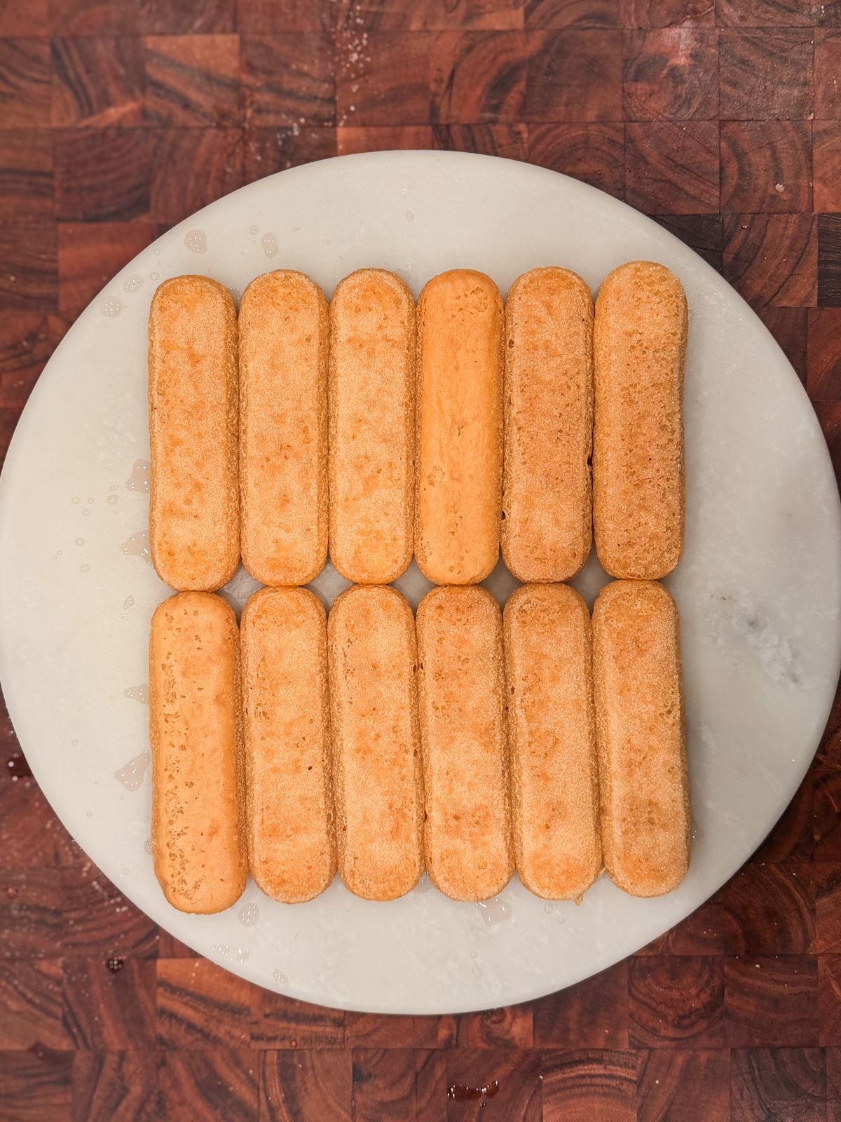 Twelve ladyfinger biscuits are neatly arranged in three rows on a round, white marble plate, which sits on a brown, checkered wooden surface.