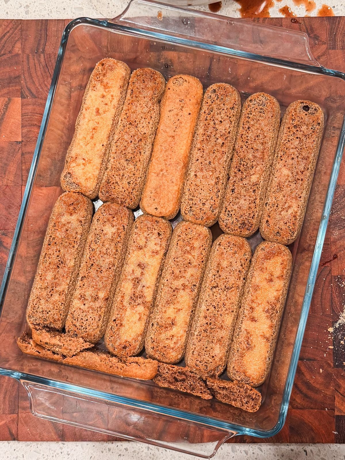 A glass baking dish with a single layer of ladyfinger cookies arranged neatly on the bottom, some slightly overlapping, on a wooden cutting board.