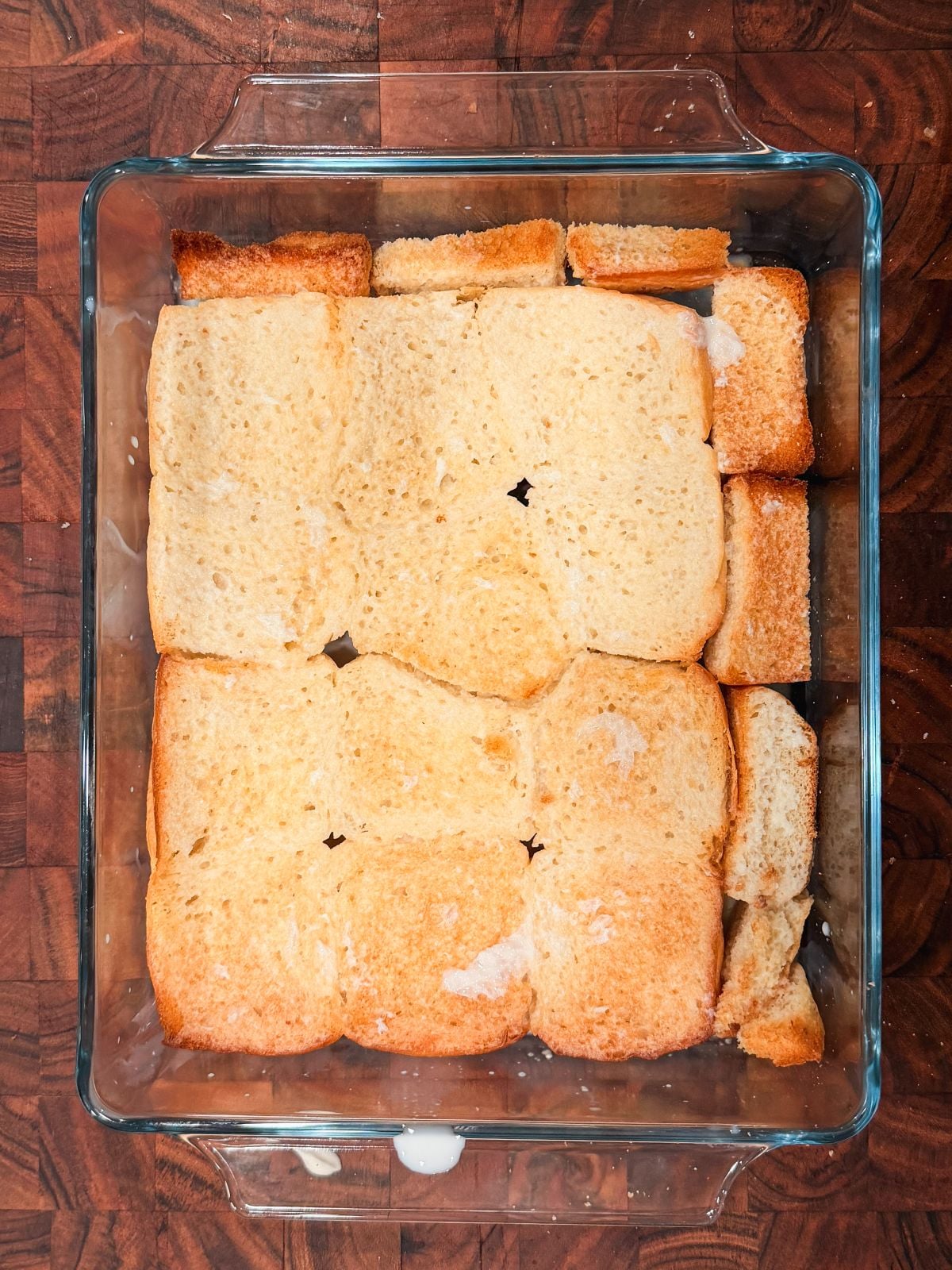 A glass baking dish filled with evenly arranged slices of bread soaked with liquid, sits on a wooden surface, ready for baking or further preparation.