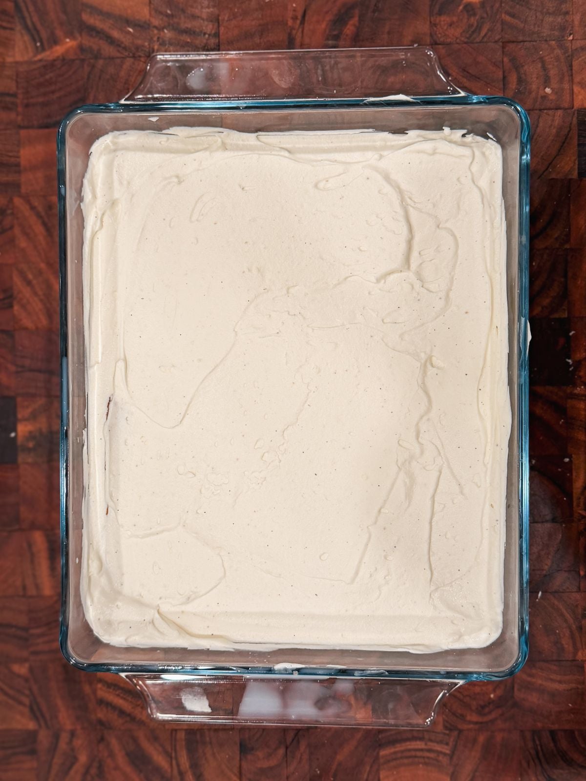 A glass baking dish filled with a smooth, creamy white mascarpone cream spread evenly over the surface, placed on a dark wooden countertop.