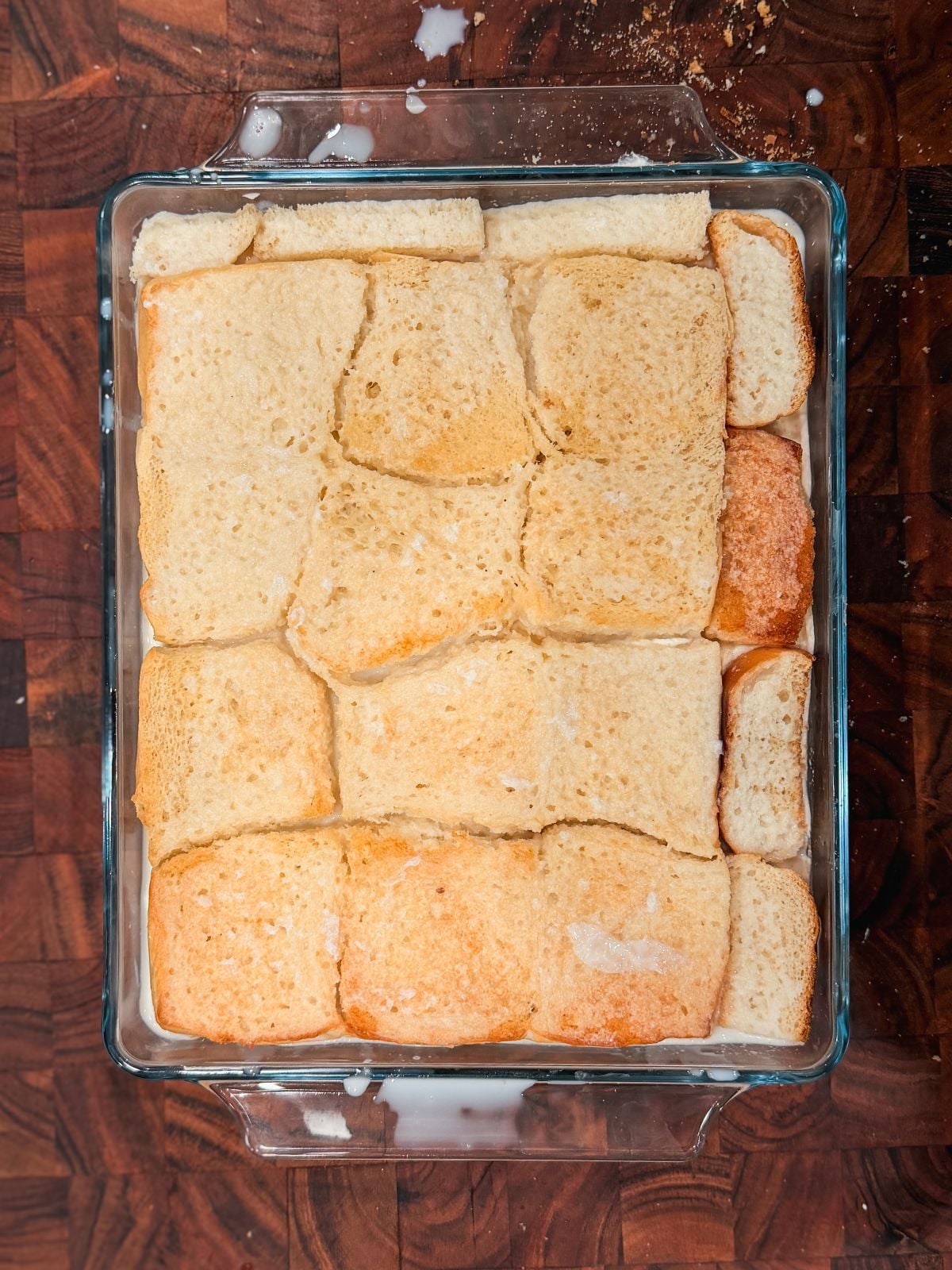 A glass baking dish filled with pieces of bread soaked in a creamy liquid, arranged in a single layer and soaked in milk, sitting on a dark wooden surface.
