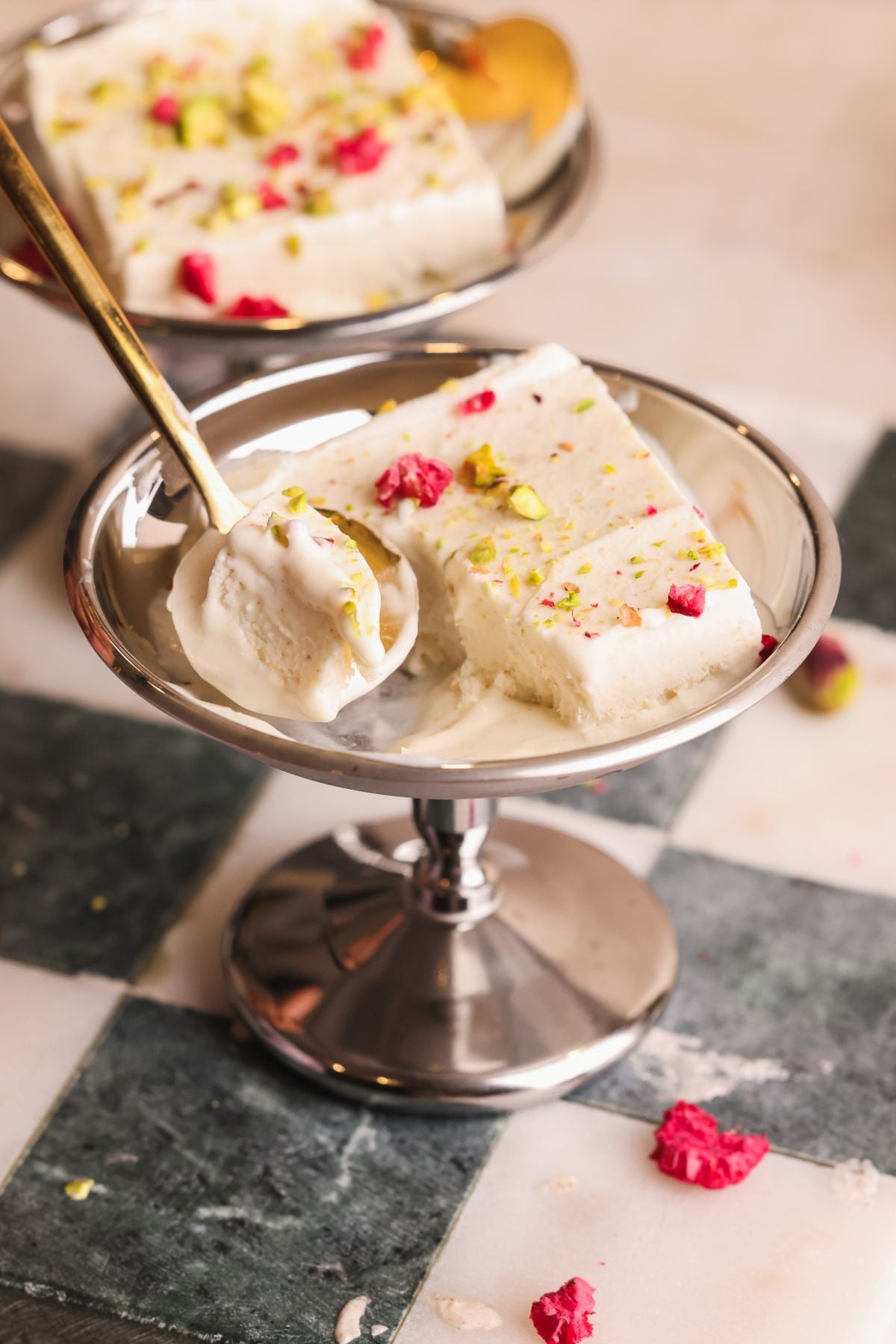 Two pieces of kulfi, an Indian frozen dessert, are served in a silver dish and garnished with chopped pistachios and dried rose petals. A spoon with a bite of kulfi rests on the edge of the dish.