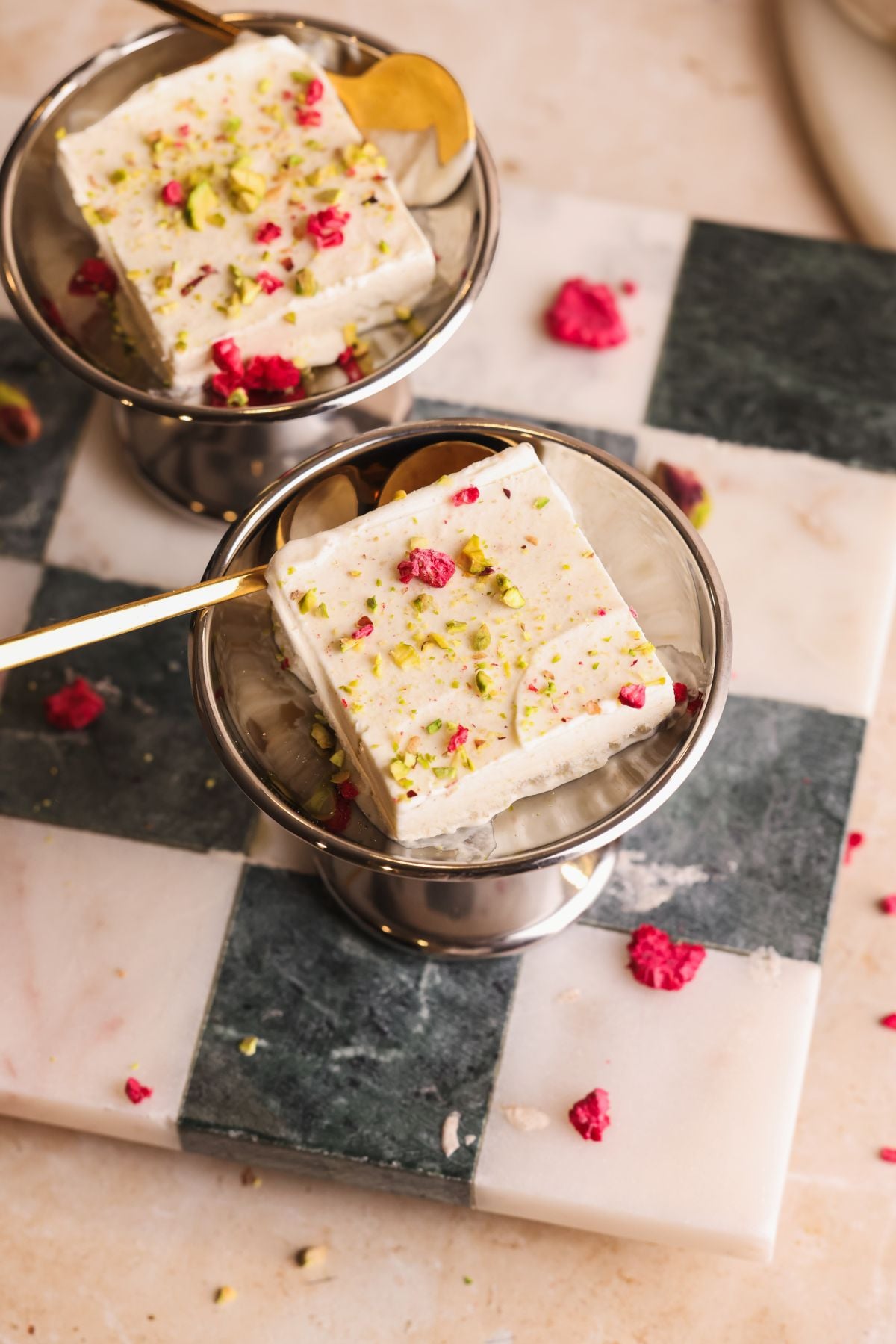 Two metal dessert cups each hold a square piece of kulfi topped with crushed pistachios and dried rose petals, placed on a checkered marble surface with scattered toppings.