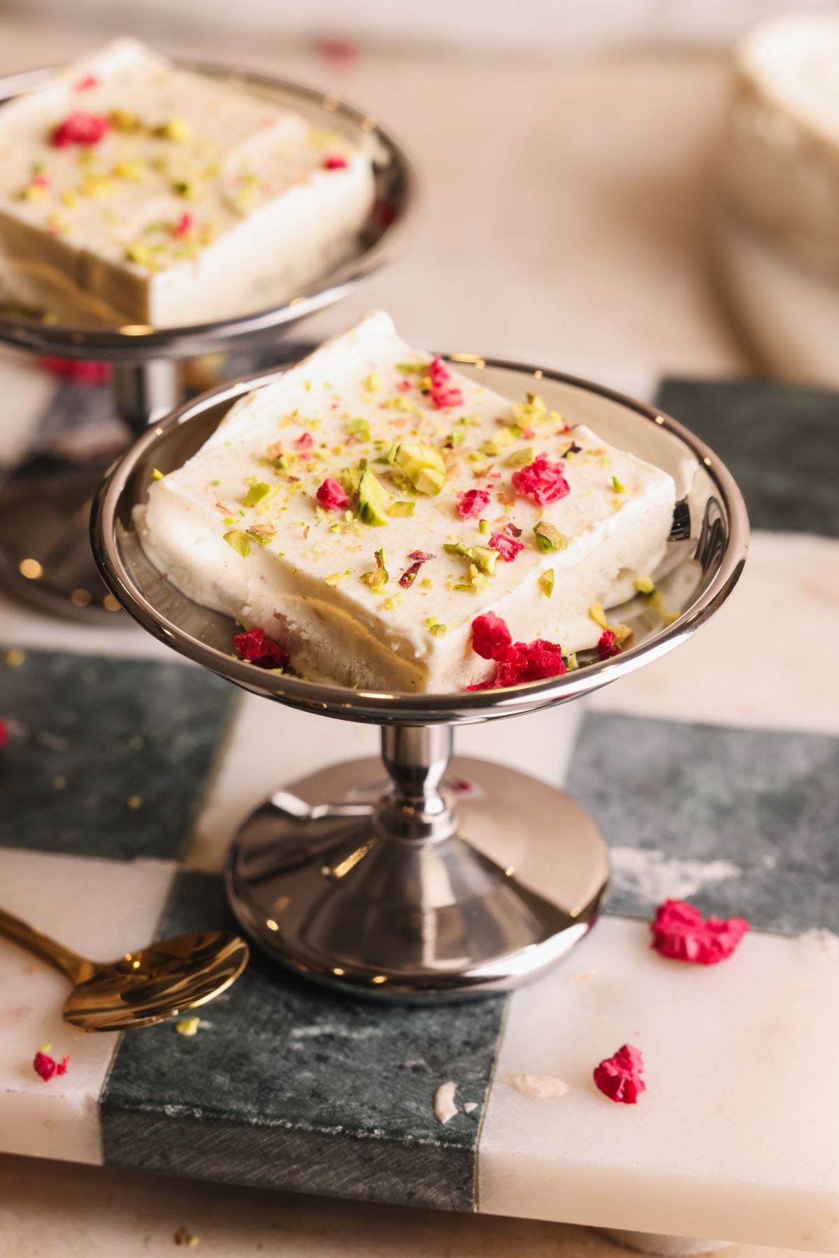 A square piece of white Indian sweet, garnished with chopped pistachios and dried rose petals, served on a small silver dessert stand placed on a black and white marble surface.