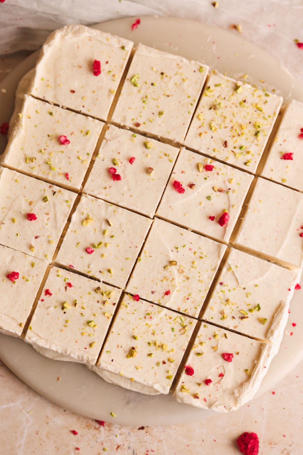Twelve square pieces of kulfi topped with crushed pistachios and bits of freeze-dried raspberries are arranged neatly on a round plate, with some crumbs scattered around.