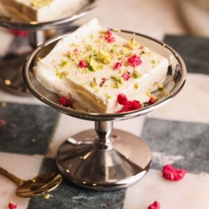 A square piece of kulfi, a South Indian ice cream dessert, garnished with chopped pistachios and dried rose petals, served in a silver dish on a checkered marble surface.