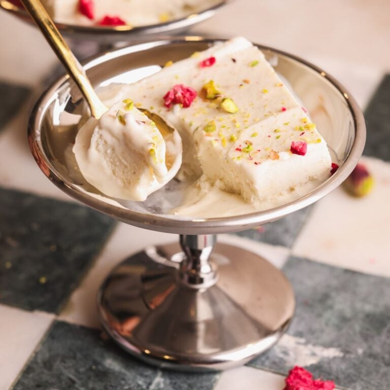 A metal dessert dish holds a slice of kulfi, an Indian ice cream, garnished with chopped pistachios and dried rose petals. A spoon with a bite-sized piece rests beside it on the dish.