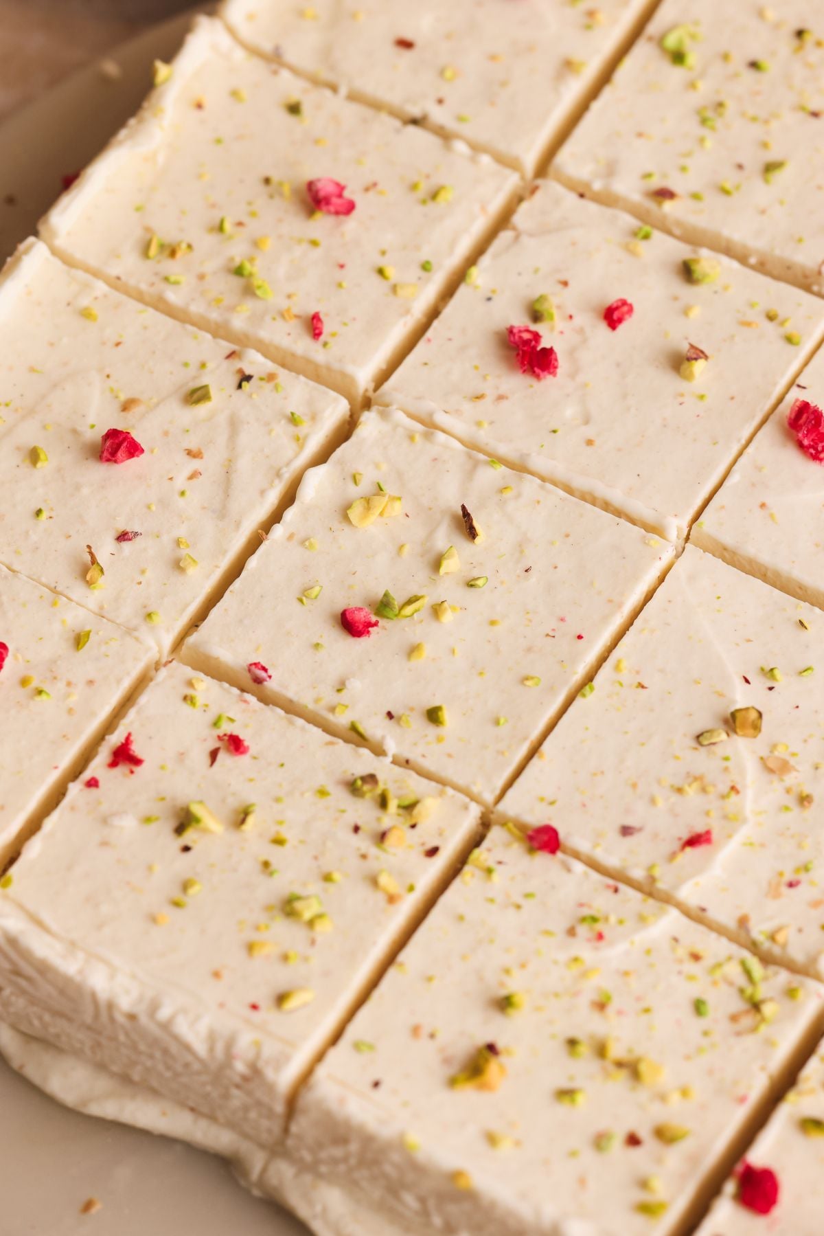 Close-up of sliced squares of creamy white kulfi, sprinkled with chopped pistachios and small bits of red topping, arranged on a plate.