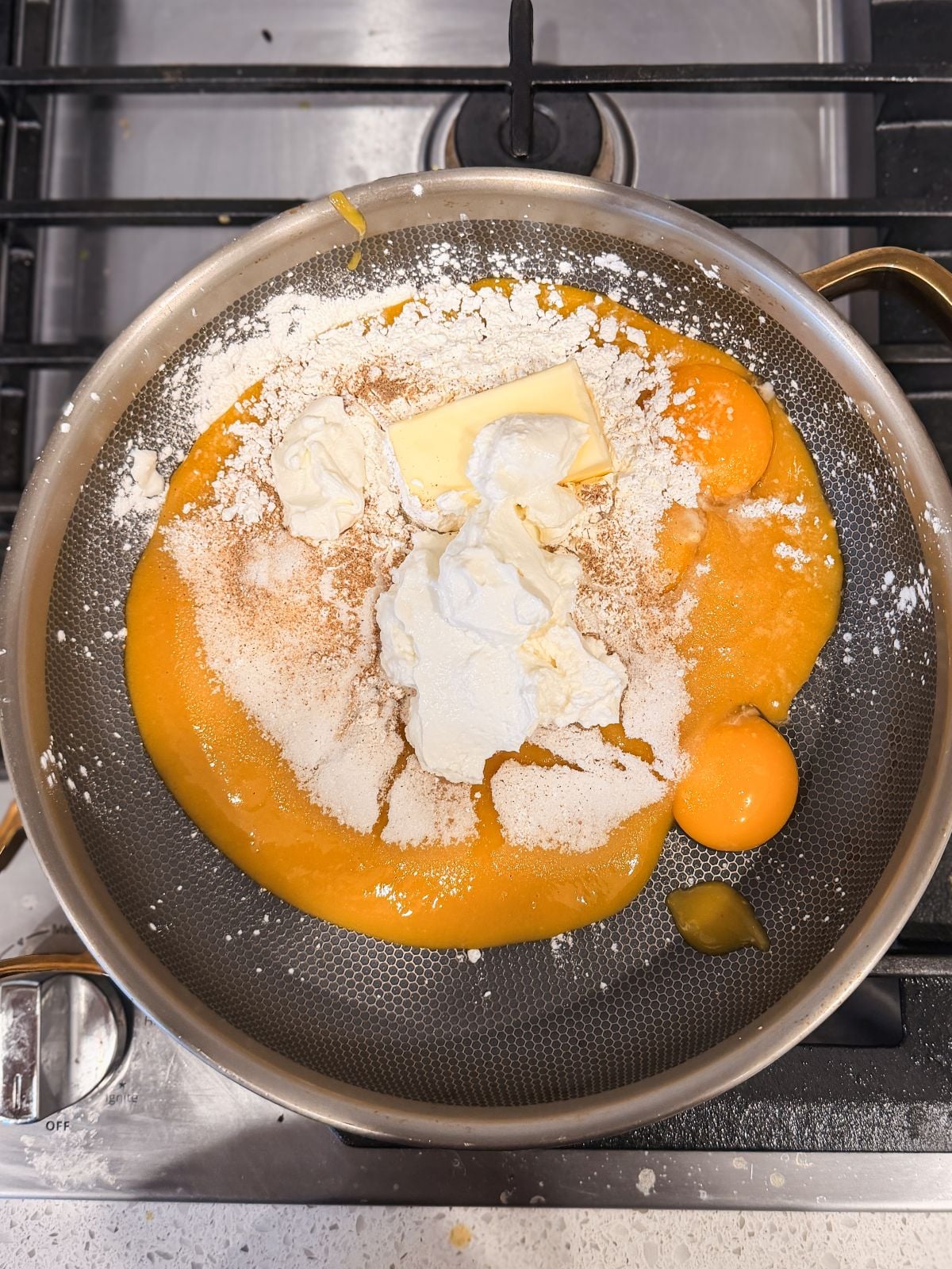 A frying pan on a stove contains eggs, flour, mango pulp, butter, and sugar, ready to be mixed for cooking. The ingredients are unmixed and sit atop a golden batter.