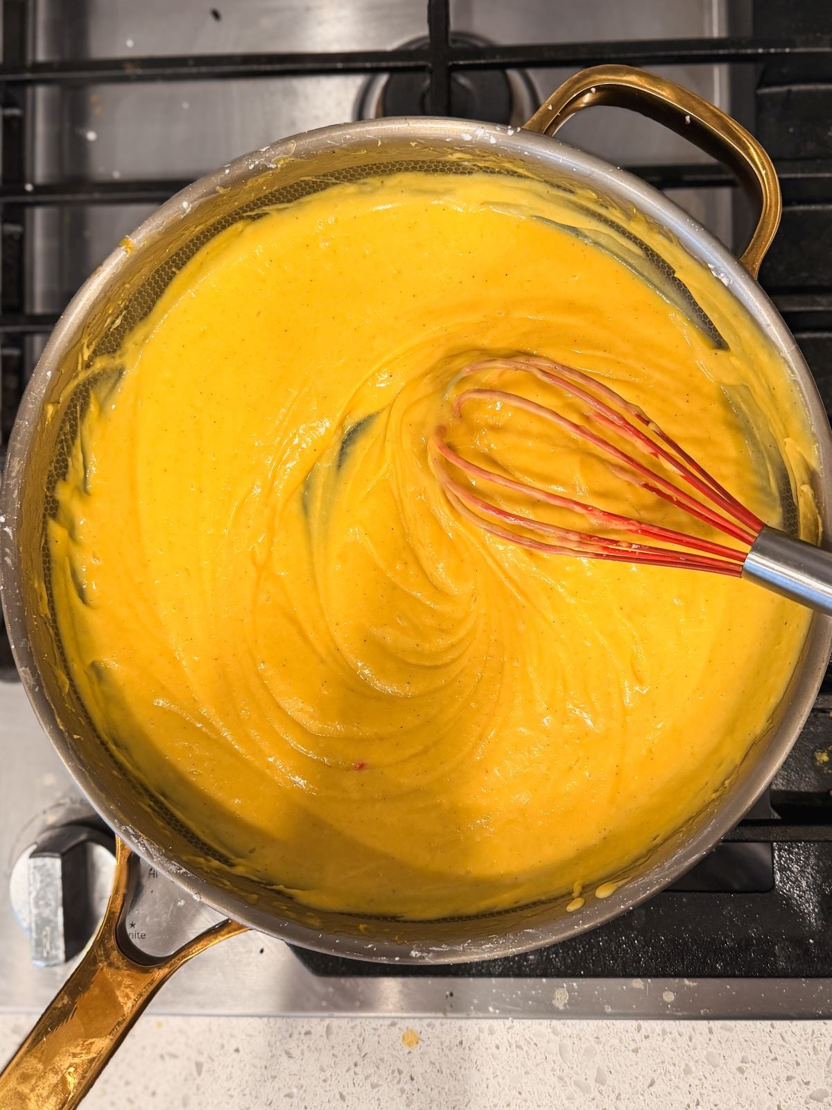 A saucepan filled with creamy yellow mango curd being whisked by a red and silver whisk, sitting on a stovetop burner.