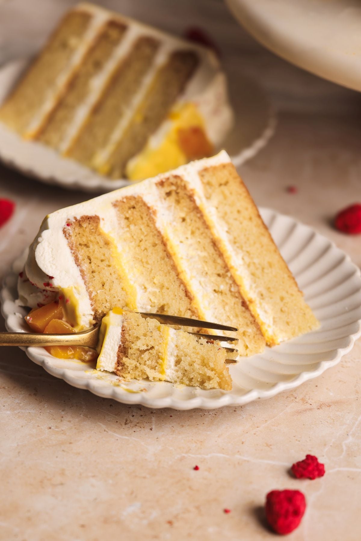 A slice of layered mango lassi cake with white frosting and mango curd filling on a white scalloped plate, with a fork taking a bite. There are a few raspberries scattered on the beige surface nearby.