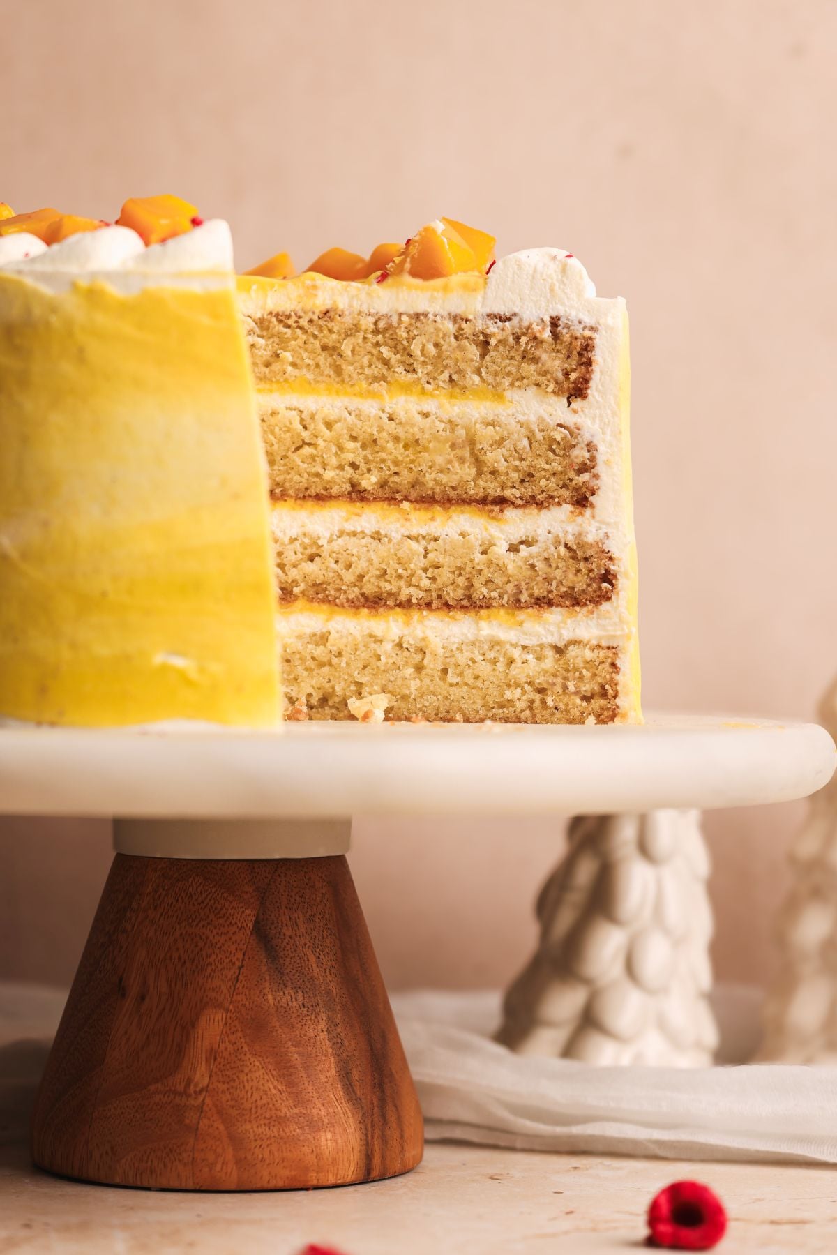 A close-up of a four-layer mango lassi cake with yellow frosting and whipped cream, topped with small mango pieces, displayed on a cake stand with a wooden base. One slice is missing, revealing the cake’s texture.