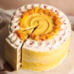 A round mango lassi cake with yellow and white frosting, decorated with whipped cream and diced mango pieces on top. One slice is being lifted from the cake.
