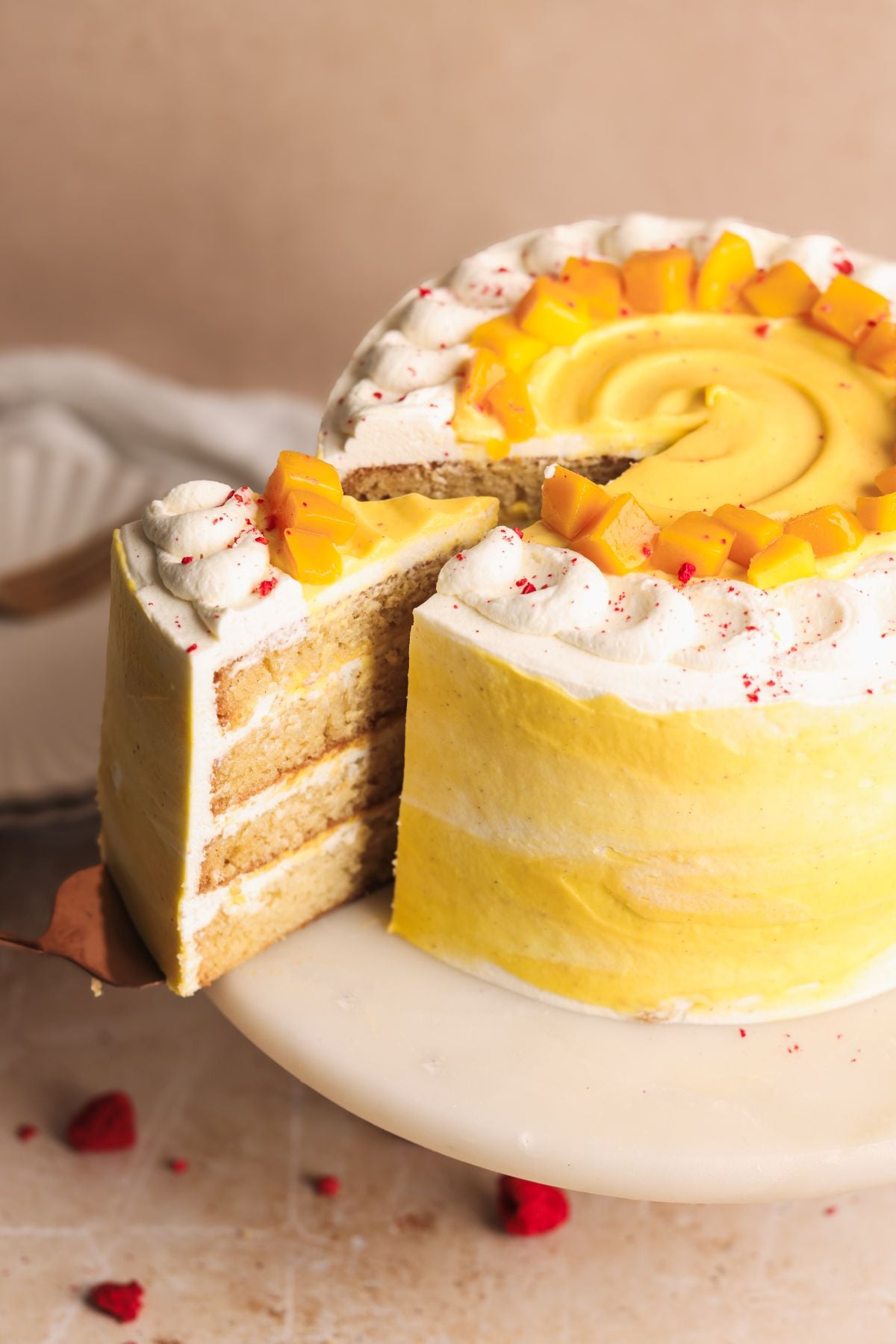 A four-layer mango lassi cake with yellow and white frosting, topped with whipped cream and diced mango, sits on a white cake stand. One slice has been cut and pulled slightly out, revealing the cake’s texture.