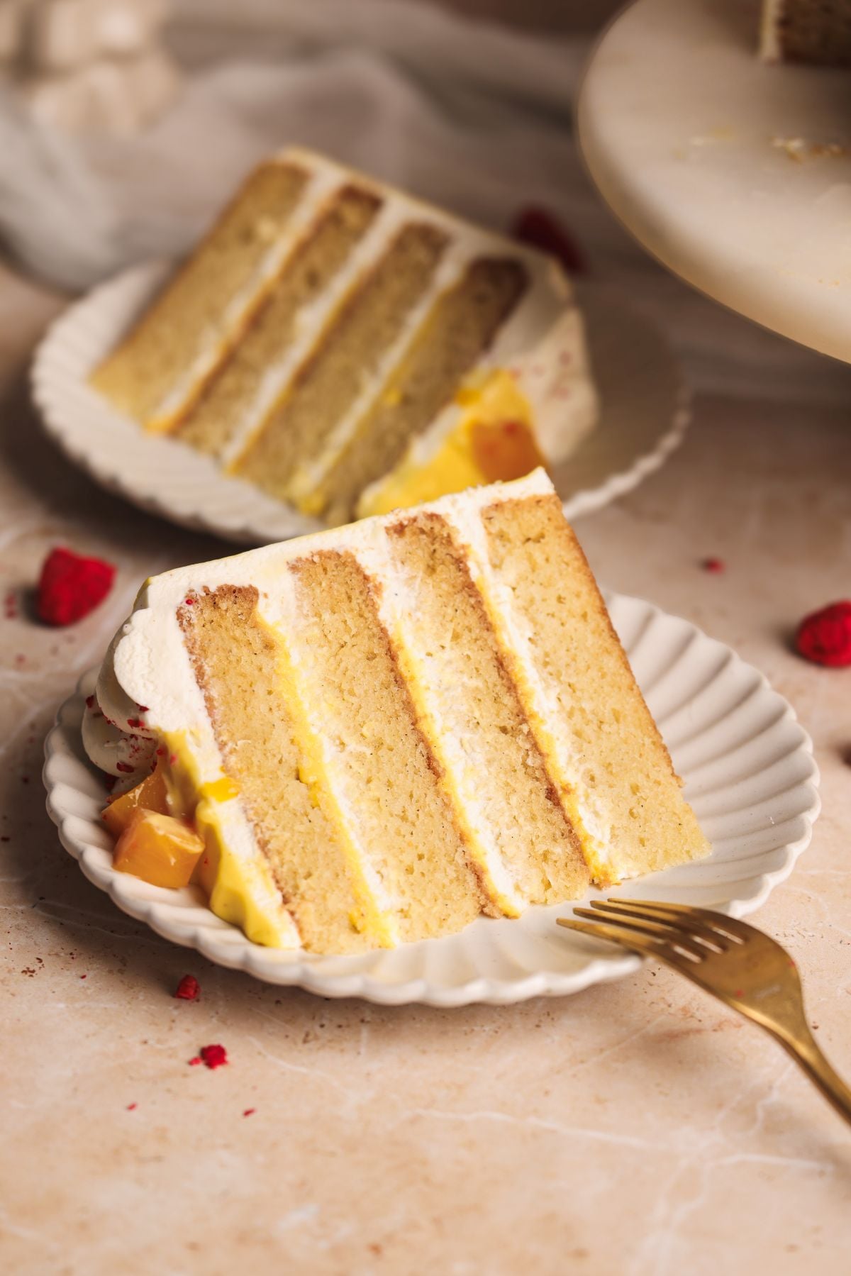 A slice of layered mango lassi cake with cream and mango curd filling on a white scalloped plate, with a fork beside it. Another slice of cake is visible in the background.