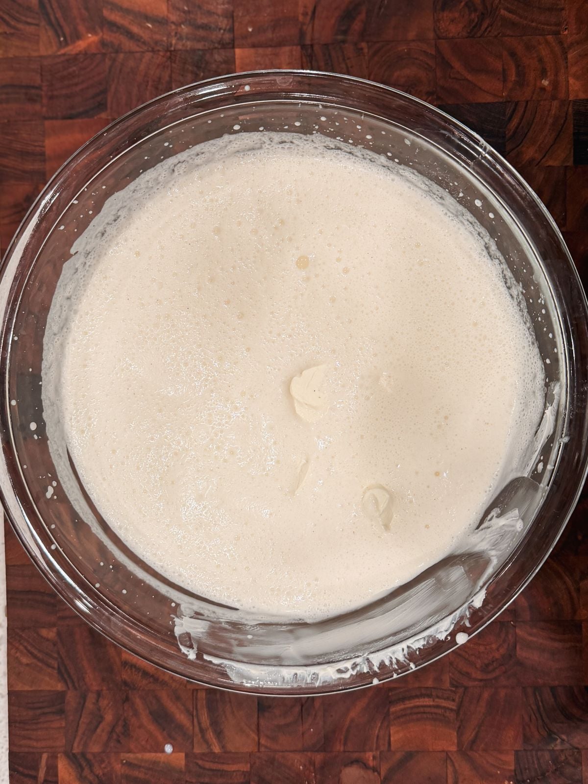A glass bowl filled with a light, frothy, cream-colored mixture sits on a dark wooden surface. Some mixture is spread on the bowl's sides, suggesting it was recently stirred or whipped. Dollop of mascarpone cheese added