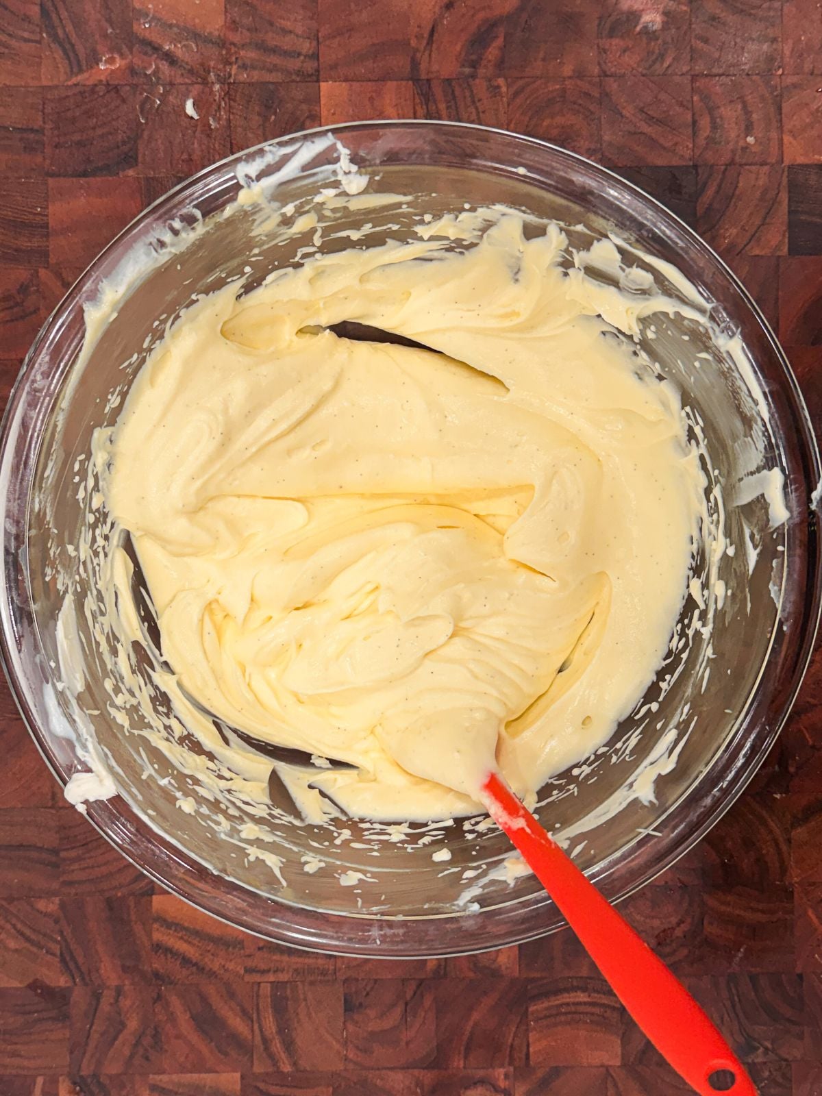A glass bowl filled with creamy, pale mascarpone cream sits on a wooden surface. A red spatula rests in the mixture, which has been partially stirred, leaving swirls and traces along the bowl's sides.