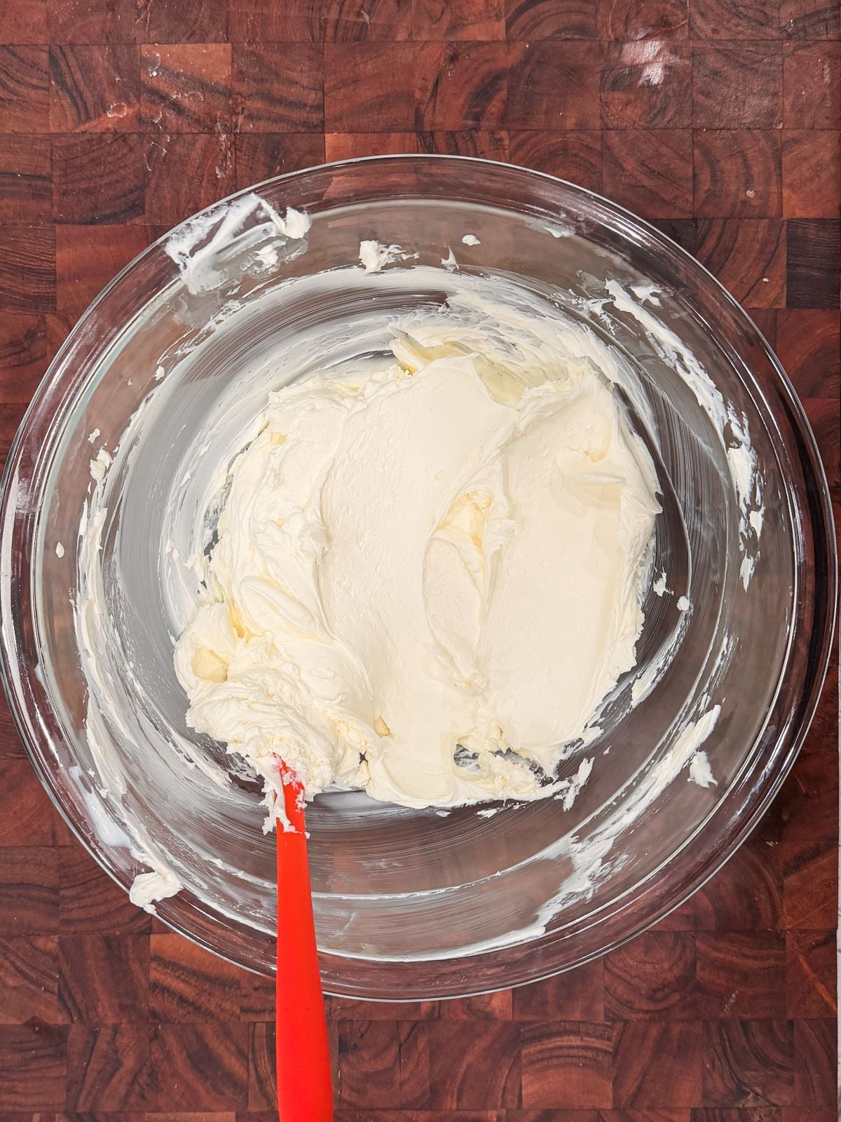 A glass mixing bowl with creamy white mascarpone and a red spatula, set on a wooden surface.