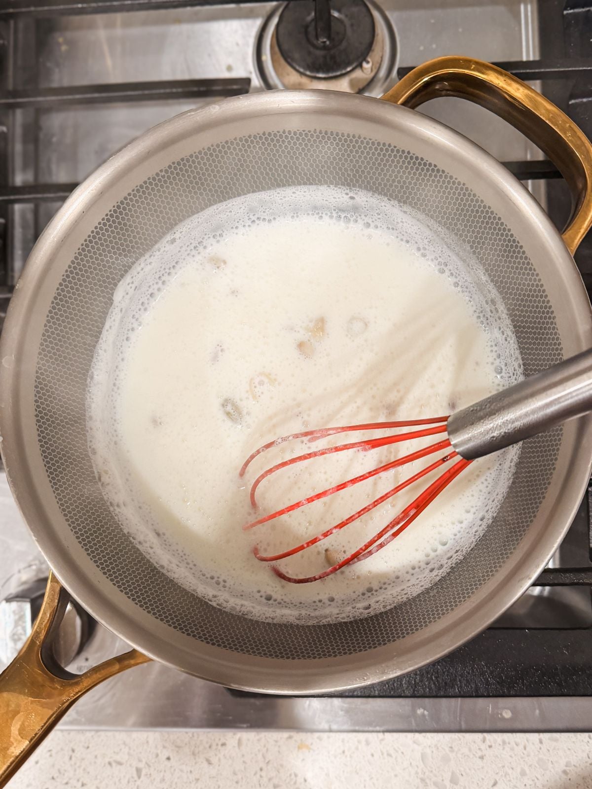 A saucepan with milk and cardamom pods is on a stove, being stirred with a red whisk. The pan has gold-colored handles and sits on a metal burner.