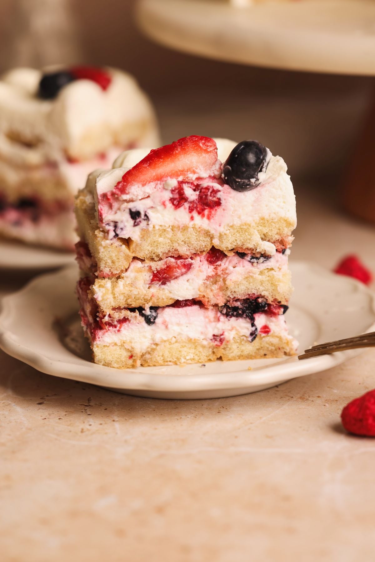 A close-up of a layered cake with cream, strawberries, and blueberries, placed on a small white plate. Another serving of the dessert is visible in the background.