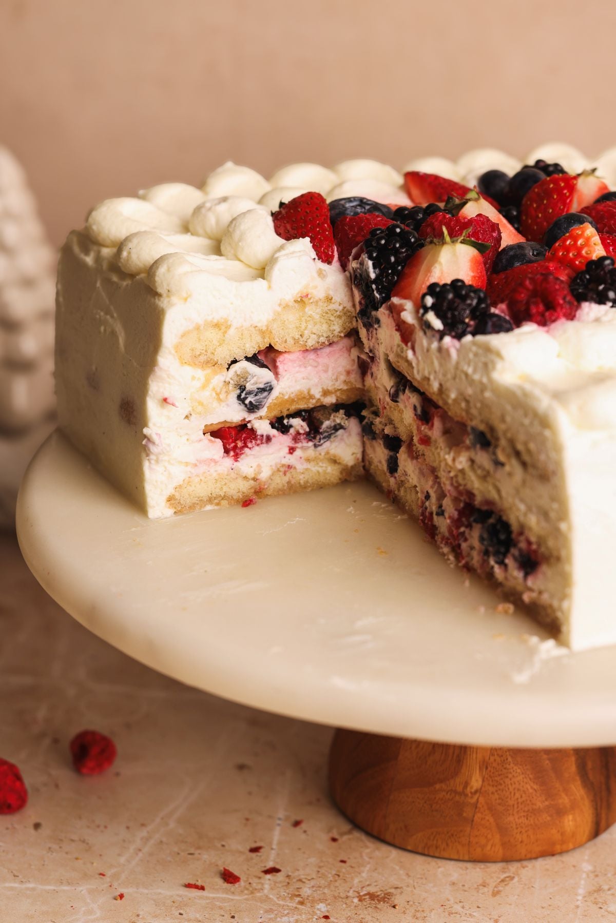 A layered vanilla cake with whipped cream and mixed berries filling, topped with assorted fresh berries and dollops of whipped cream, sits on a round cake stand with a slice removed.