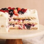 A sliced cake on a stand, showing layers of whipped cream, ladyfingers, and mixed berries like strawberries, raspberries, and blackberries. The top is decorated with more cream and fresh berries.