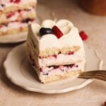 A slice of layered dessert with creamy filling and mixed berries, topped with a swirl of cream, a blueberry, and a strawberry, served on a beige plate with a fork beside it.
