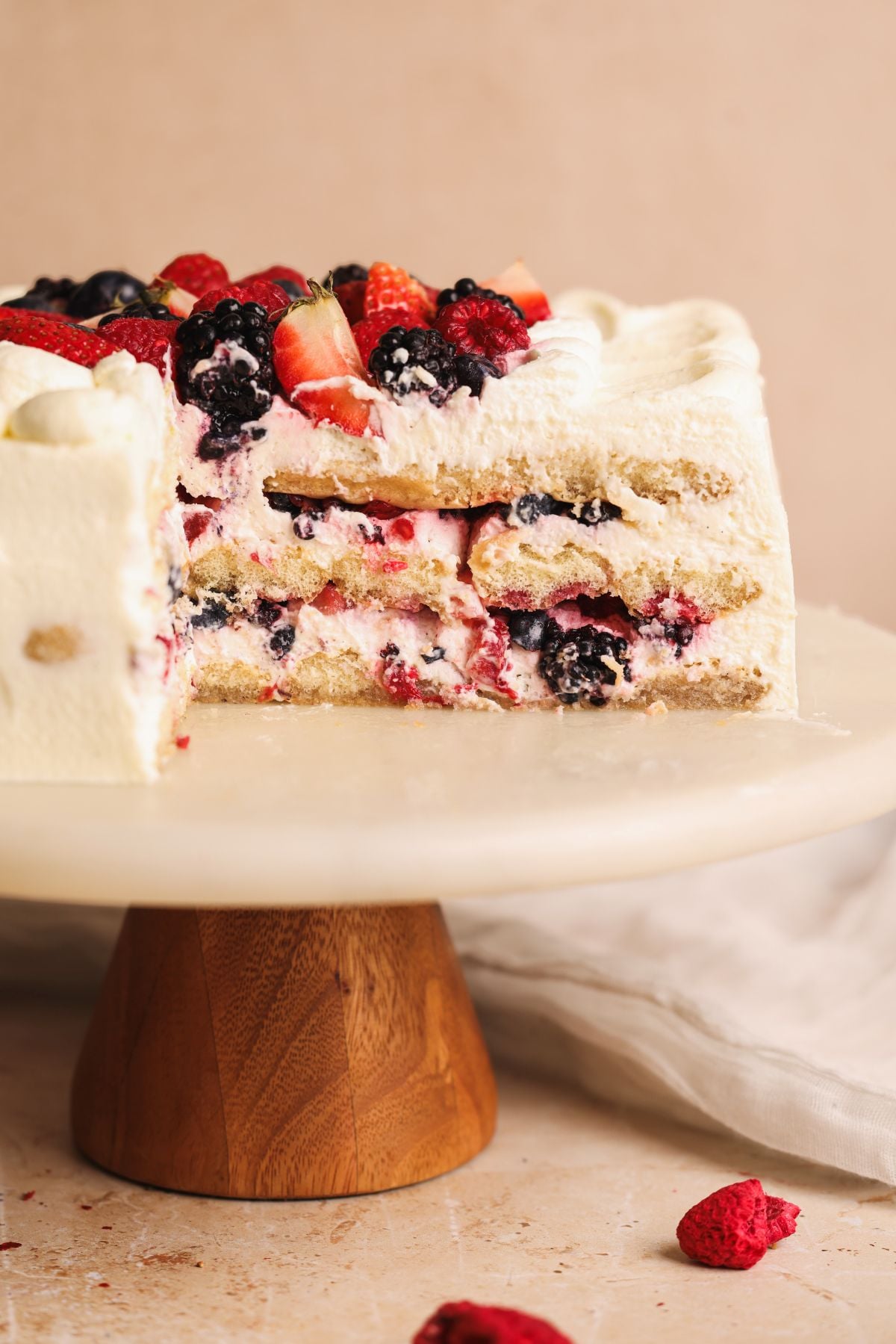 A layered cake with white frosting and mixed berries, including strawberries, raspberries, and blackberries, on top and inside. A large slice is cut out, revealing the berry and cream filling. The cake sits on a round marble stand.