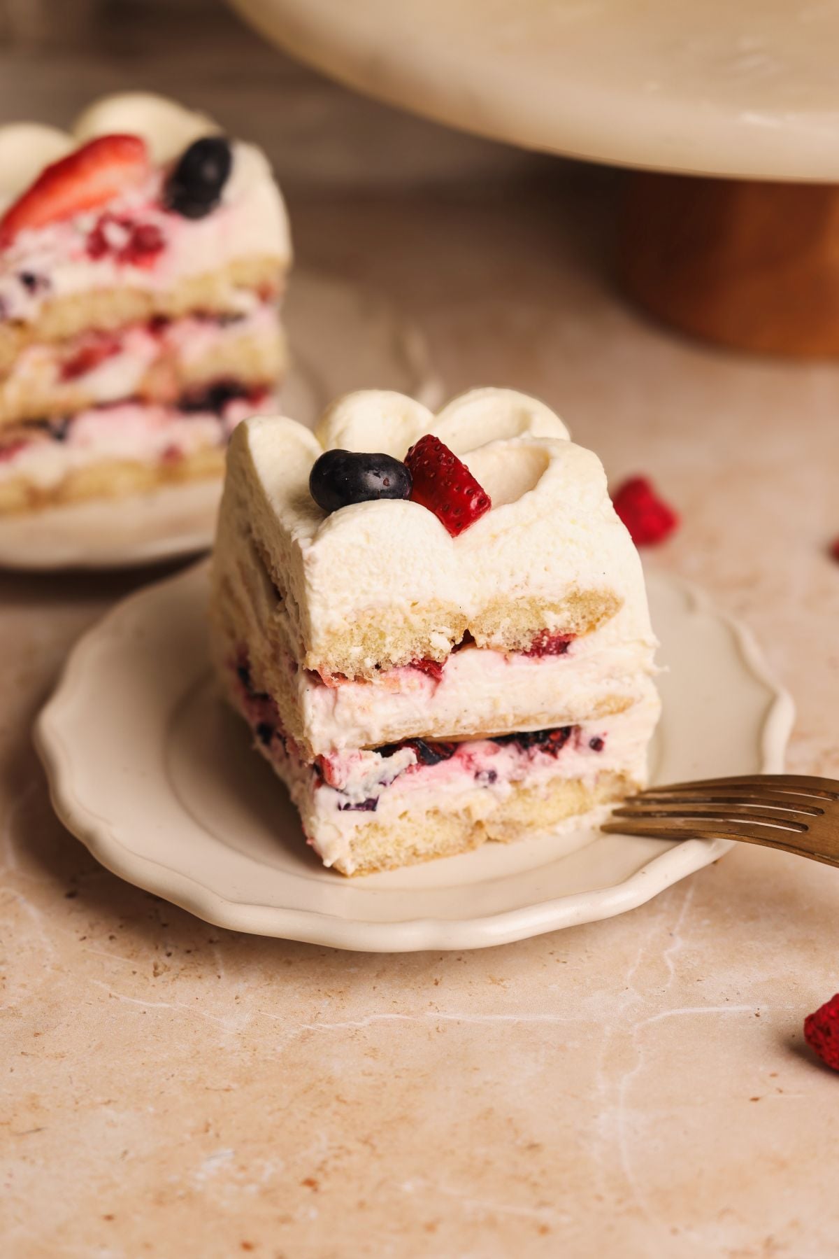 A slice of layered cake with cream, berries, and sponge, topped with a strawberry and blueberry, sits on a small plate with a fork. Another slice is in the background.