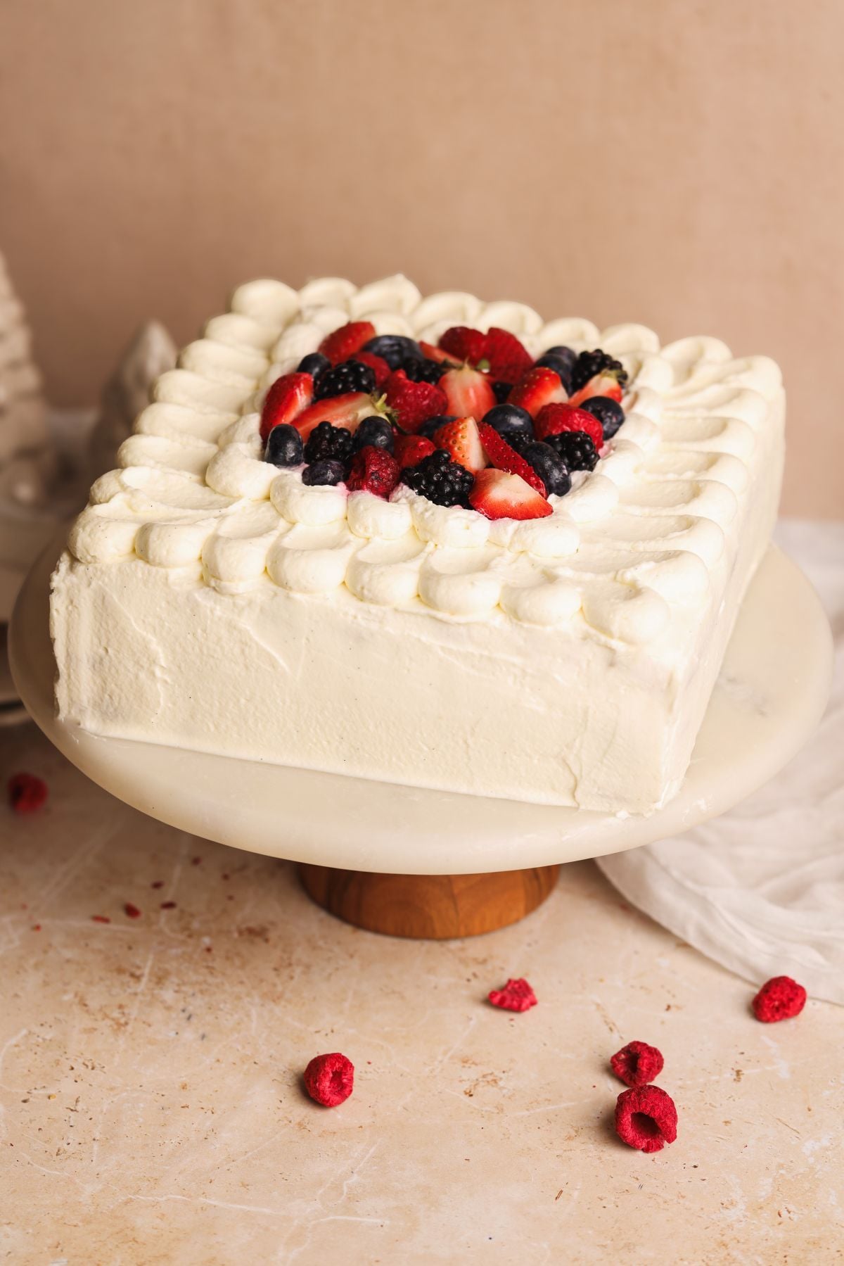 A square white frosted cake topped with strawberries, blackberries, raspberries, and blueberries, displayed on a marble cake stand with a few raspberries scattered on the surface below.