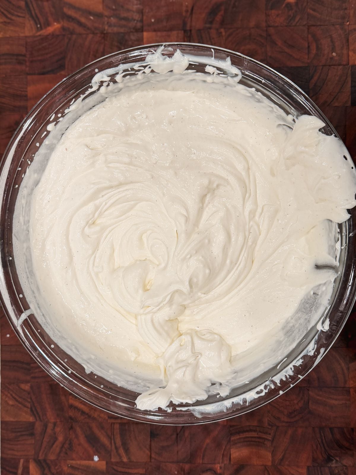 A glass bowl filled with smooth, fluffy whipped mascarpone cream sits on a dark wooden surface. The cream appears freshly whipped, with soft peaks and swirled textures visible on top.