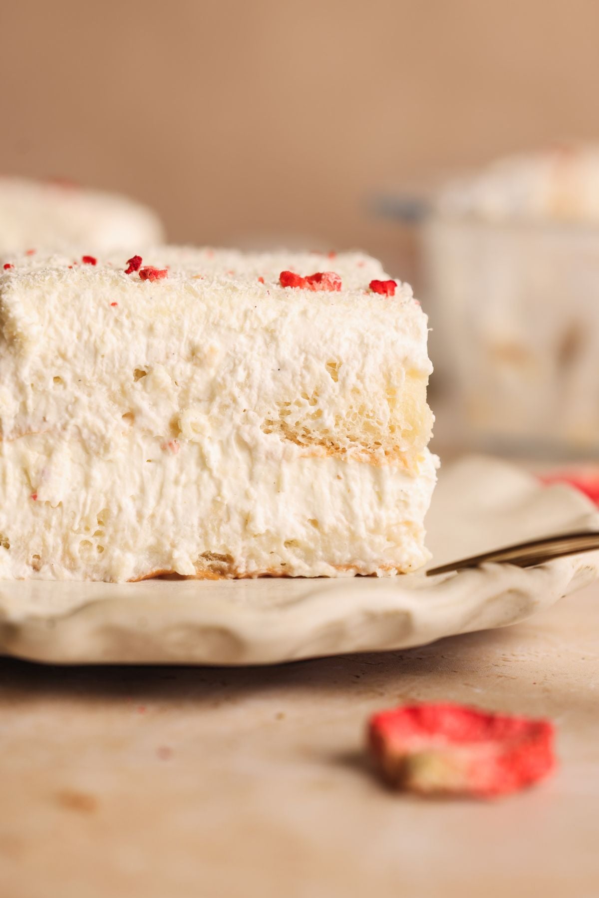 A close-up of a slice of creamy layered rus malai tiramisu, topped with whipped cream and freeze dried strawberries, served on a white, textured plate. A fork and red fruit pieces are nearby.