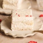 A square slice of creamy, layered rus malai tiramisu with grated white chocolate sits on a scalloped beige plate, with a gold fork beside it and another slice blurred in the background.