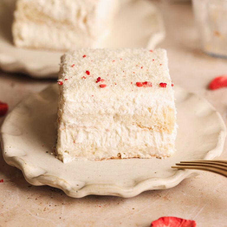 A square slice of creamy, layered rus malai tiramisu with grated white chocolate sits on a scalloped beige plate, with a gold fork beside it and another slice blurred in the background.