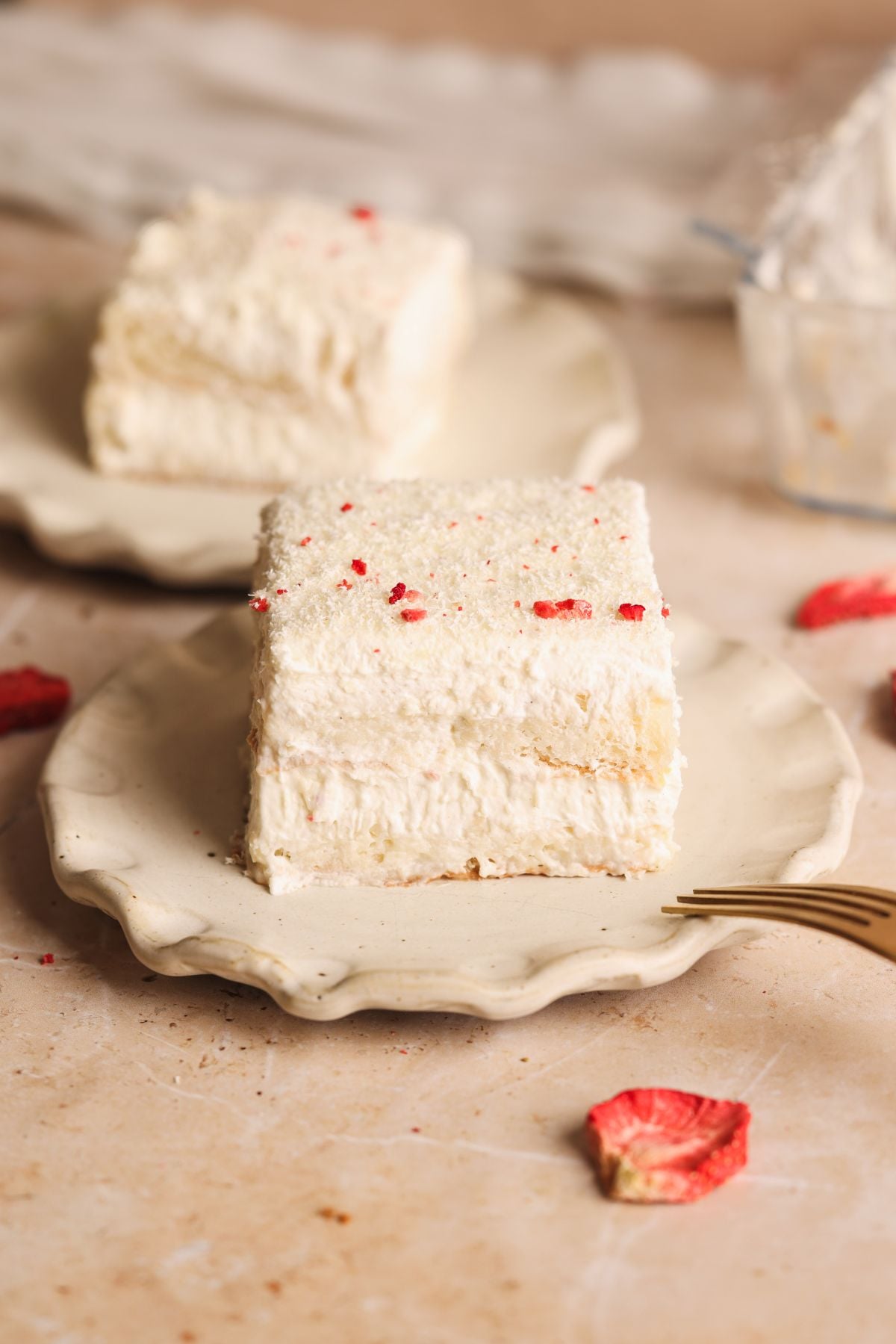 A square slice of creamy, layered rus malai tiramisu with a light, fluffy texture sits on a beige ceramic plate, garnished with a few crushed freeze dried strawberries, with another slice in the background and dried fruit pieces nearby.